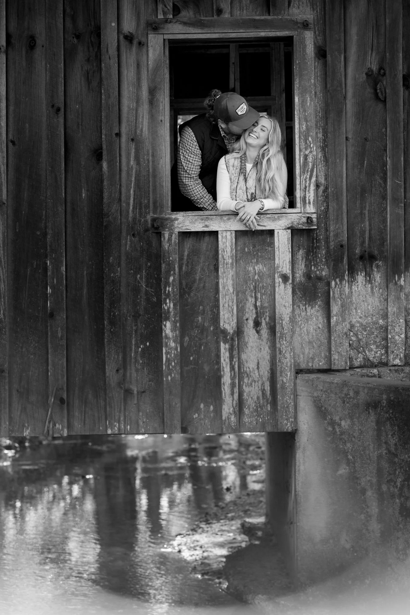 A black and white photo of a man and woman leaning out of a wooden barn window. The man is kissing the woman on her head. The barn has a rustic wooden exterior and a small creek or pond with reflection down below.