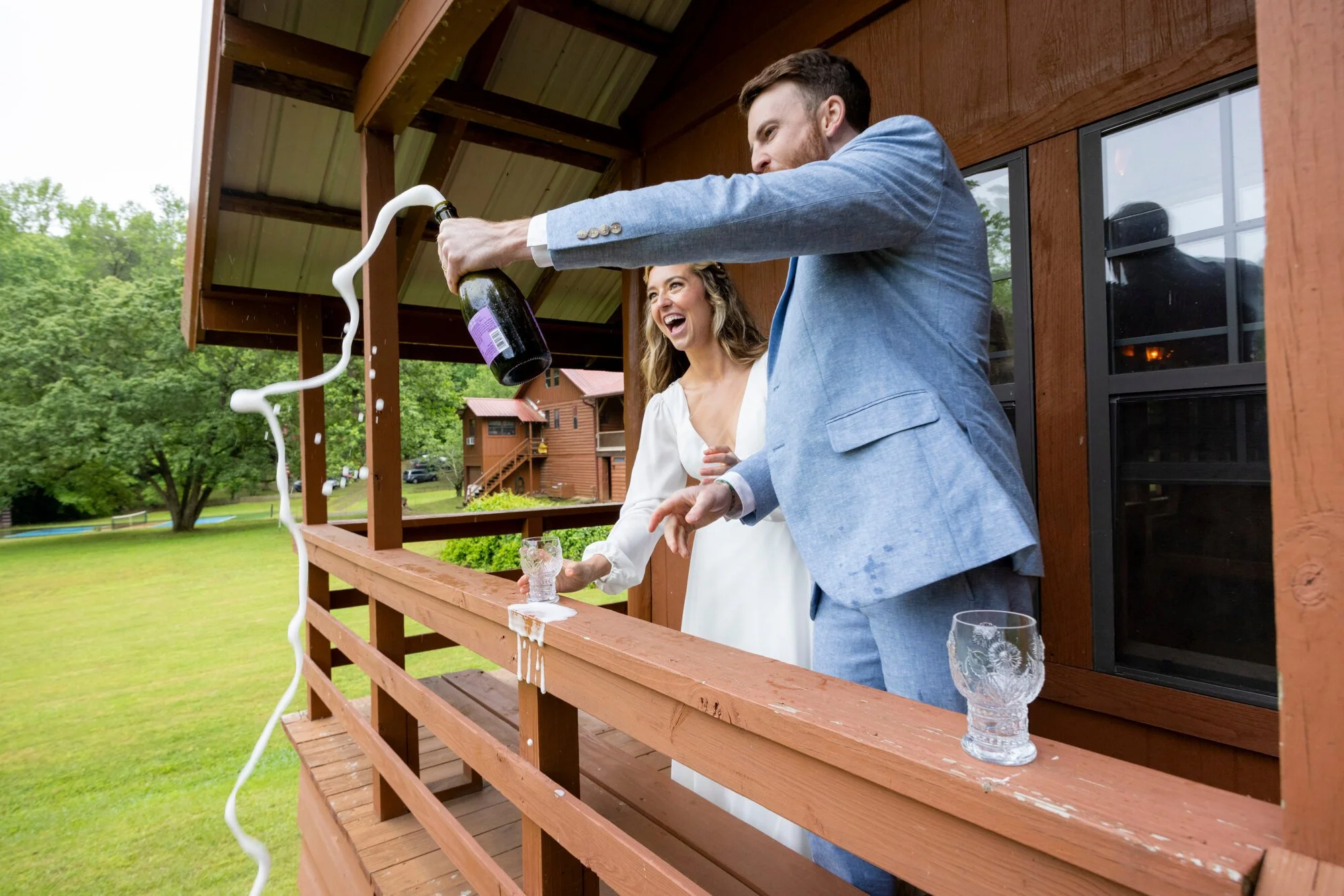 A man in a blue suit opening a bottle of champagne with a woman in a white dress smiling on a wooden porch, with spilled champagne on the floor and glasses on the railing, outdoors with trees and a building in the background.
