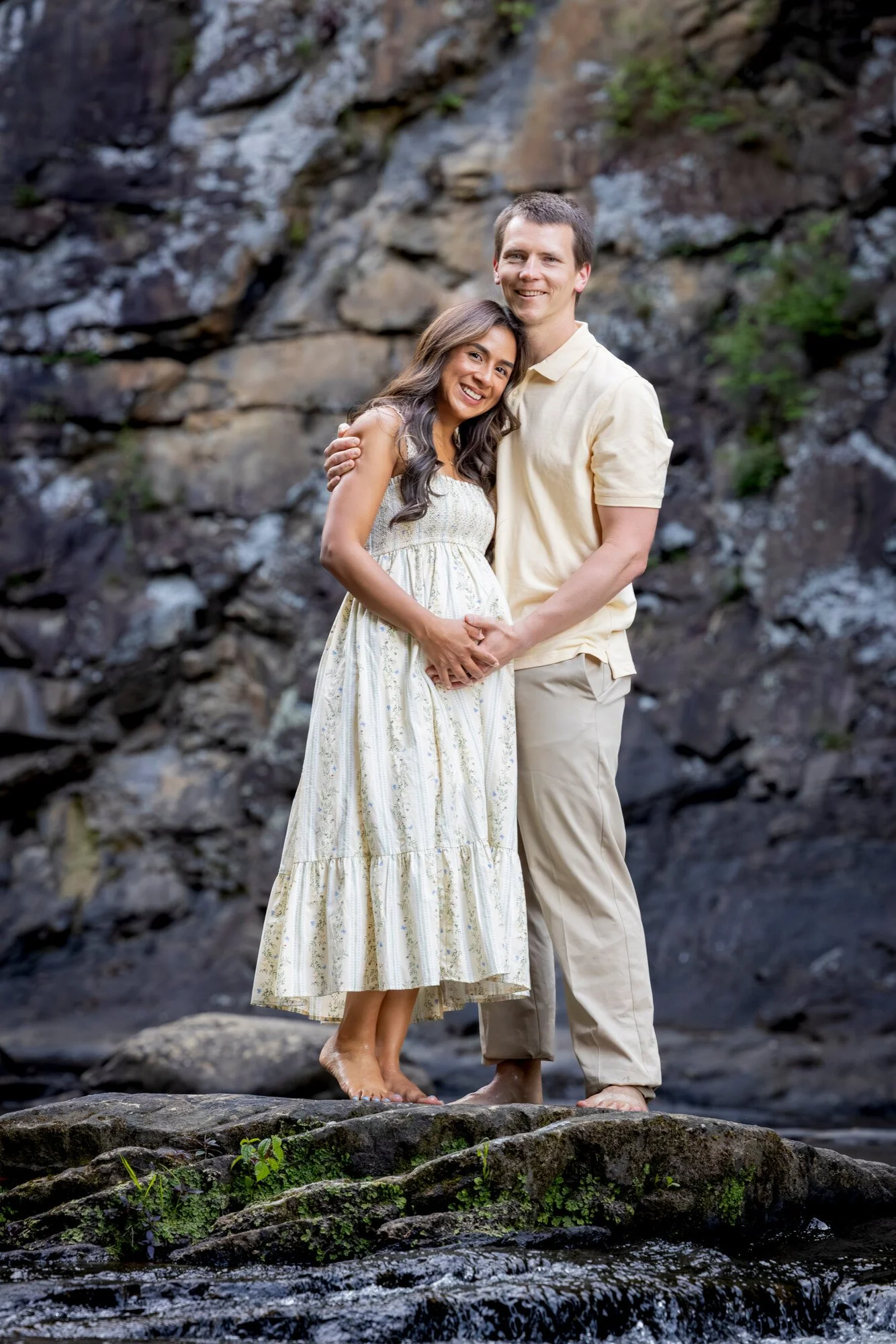 A couple standing barefoot on rocks by a creek, embracing and smiling. The woman wears a white dress, and the man is in a light-colored shirt and pants, with a rocky background and some green plants.