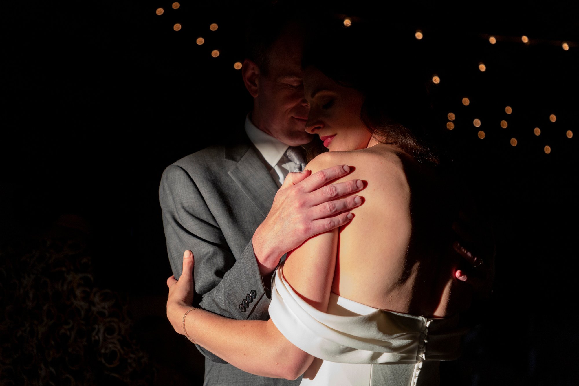 A couple dancing closely at a dimly lit event, with the man wearing a gray suit and the woman in a white dress, their faces touching and eyes closed.