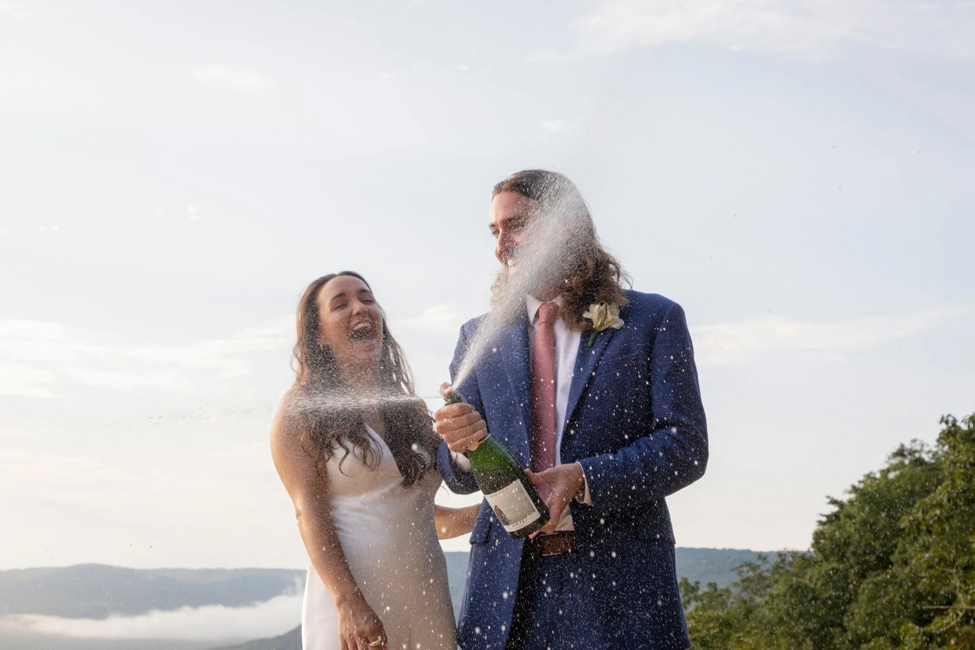 A bride and groom celebrating outdoors with the bride spraying champagne at the groom, both smiling and enjoying the moment.