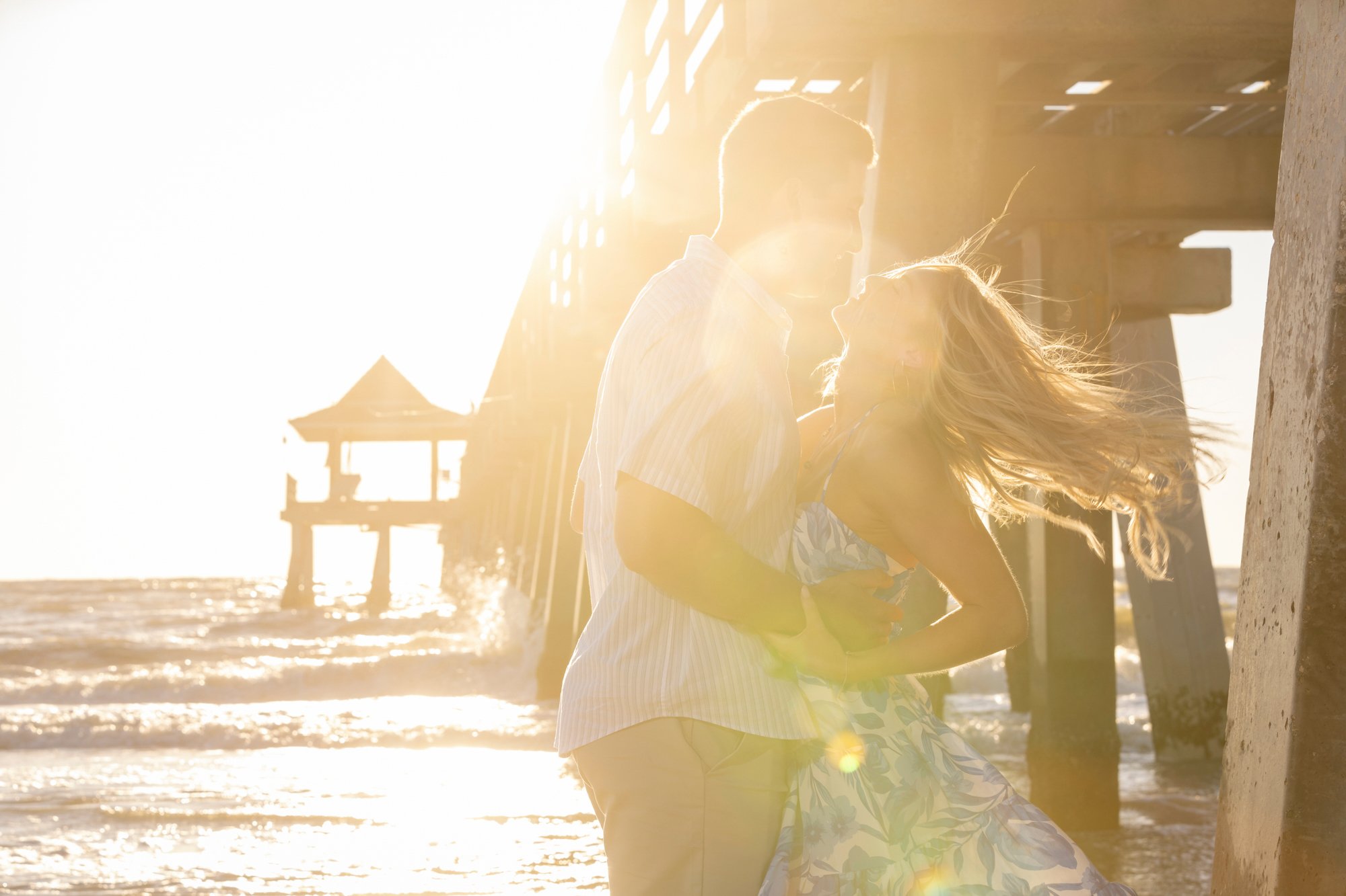A couple holding each other on a beach with a pier in the background, backlit by the setting sun.