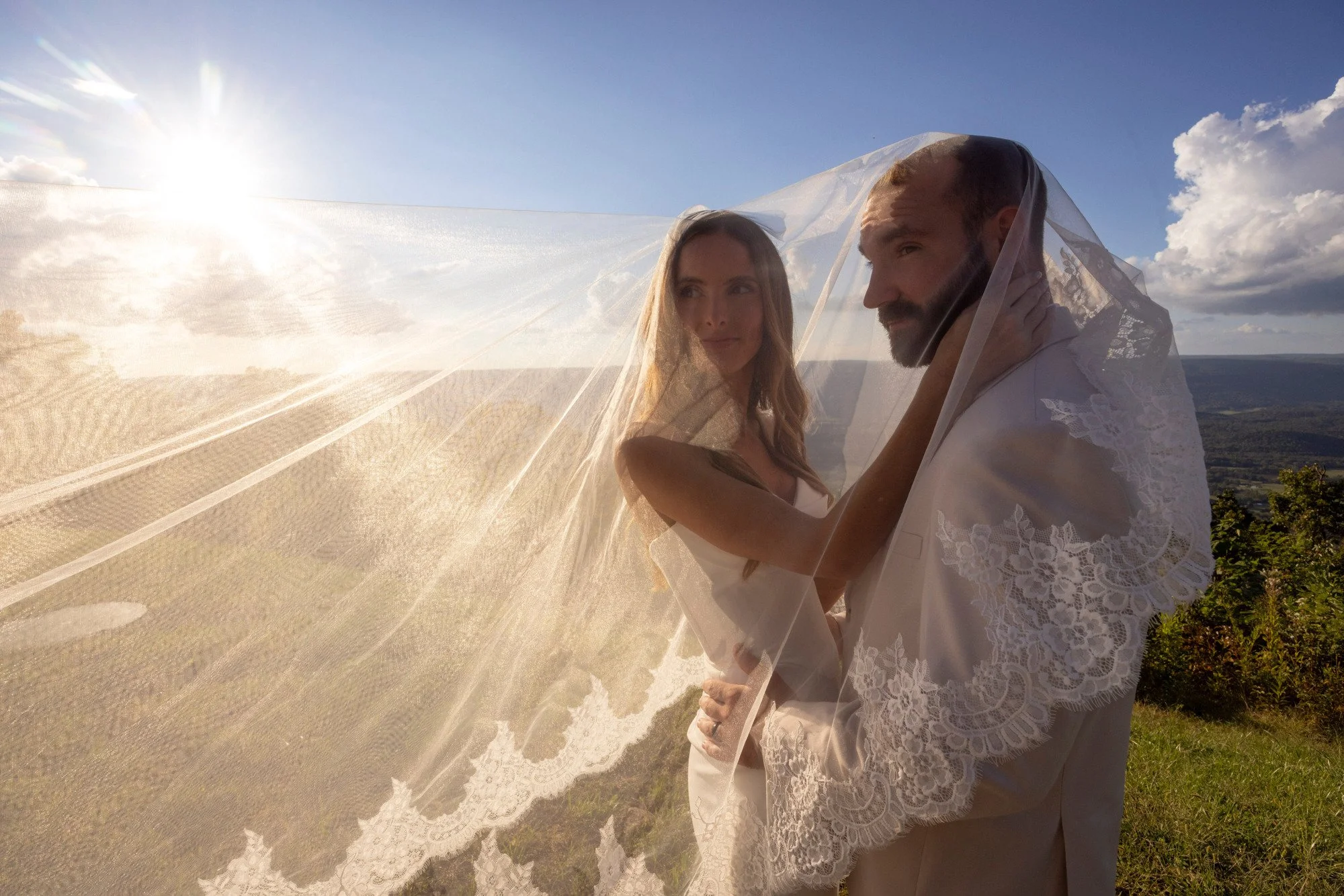 A couple dressed in wedding attire standing outdoors with a scenic landscape and blue sky in the background, holding a lace wedding veil that creates a semi-transparent overlay.
