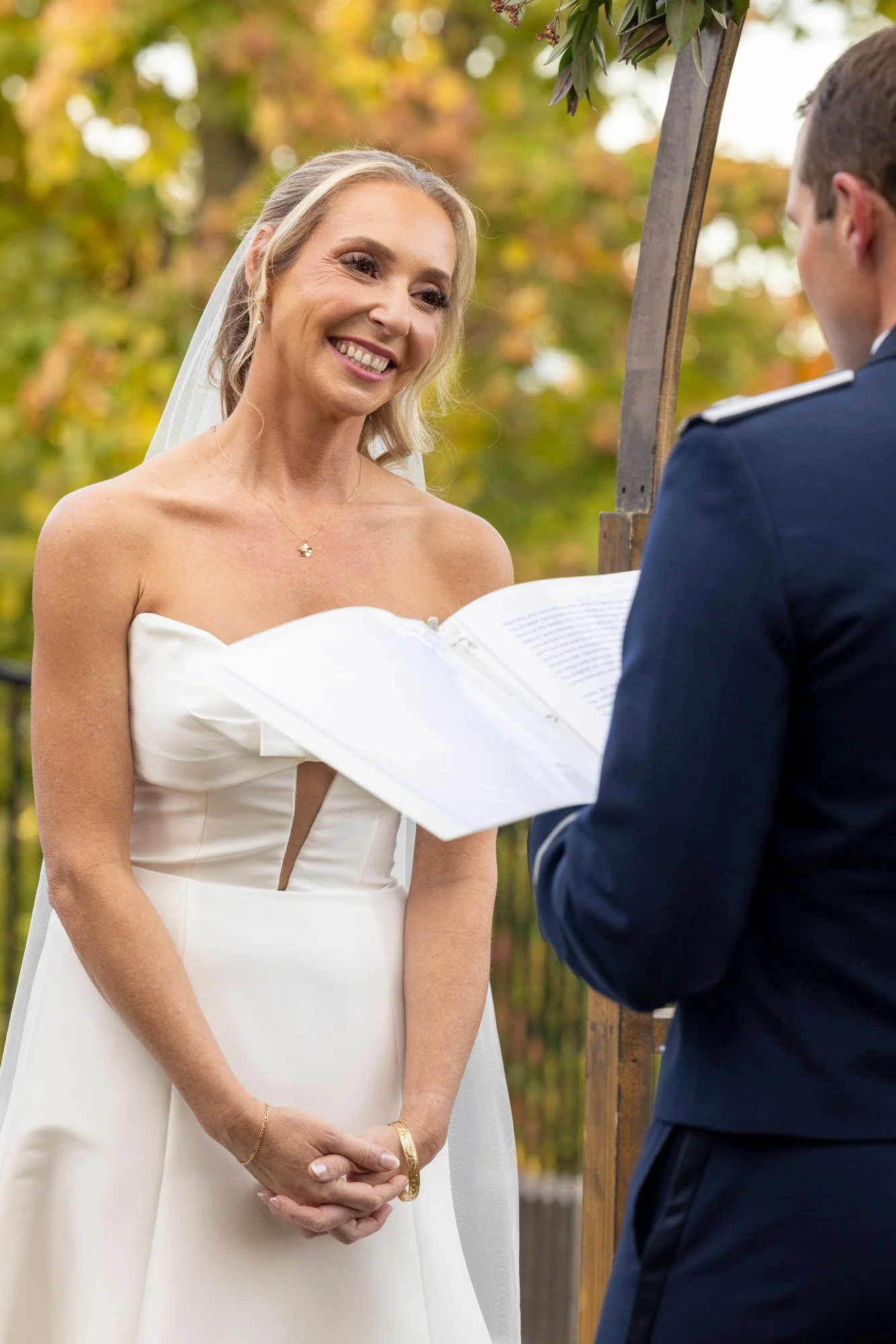 A bride in a strapless white wedding dress with a veil smiles at a groom in a dark suit during an outdoor wedding ceremony with autumn foliage in the background.