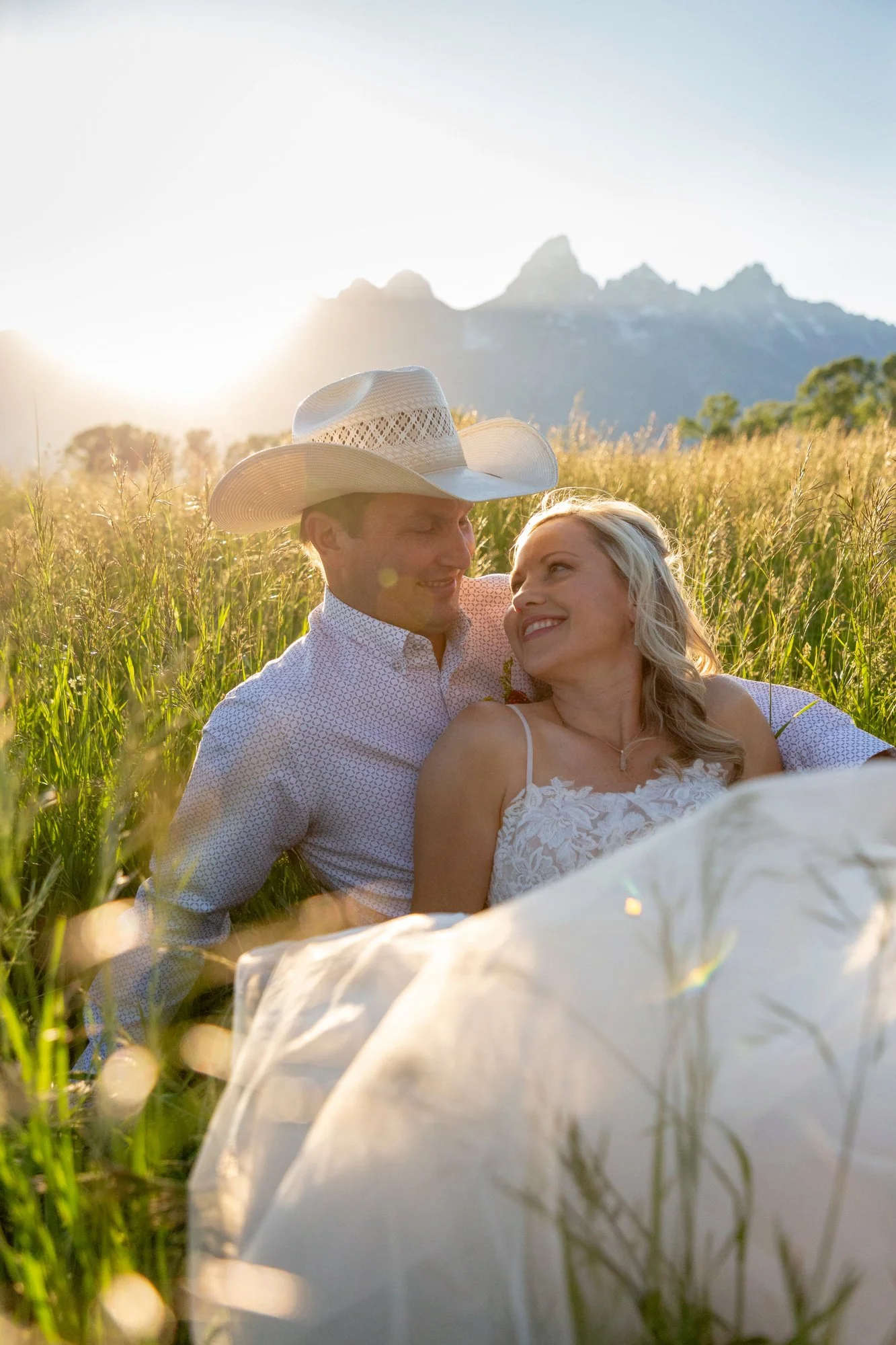 A couple relaxing in a grassy field during sunset, with mountains in the background, woman wearing a white lace dress and man wearing a cowboy hat and patterned shirt.