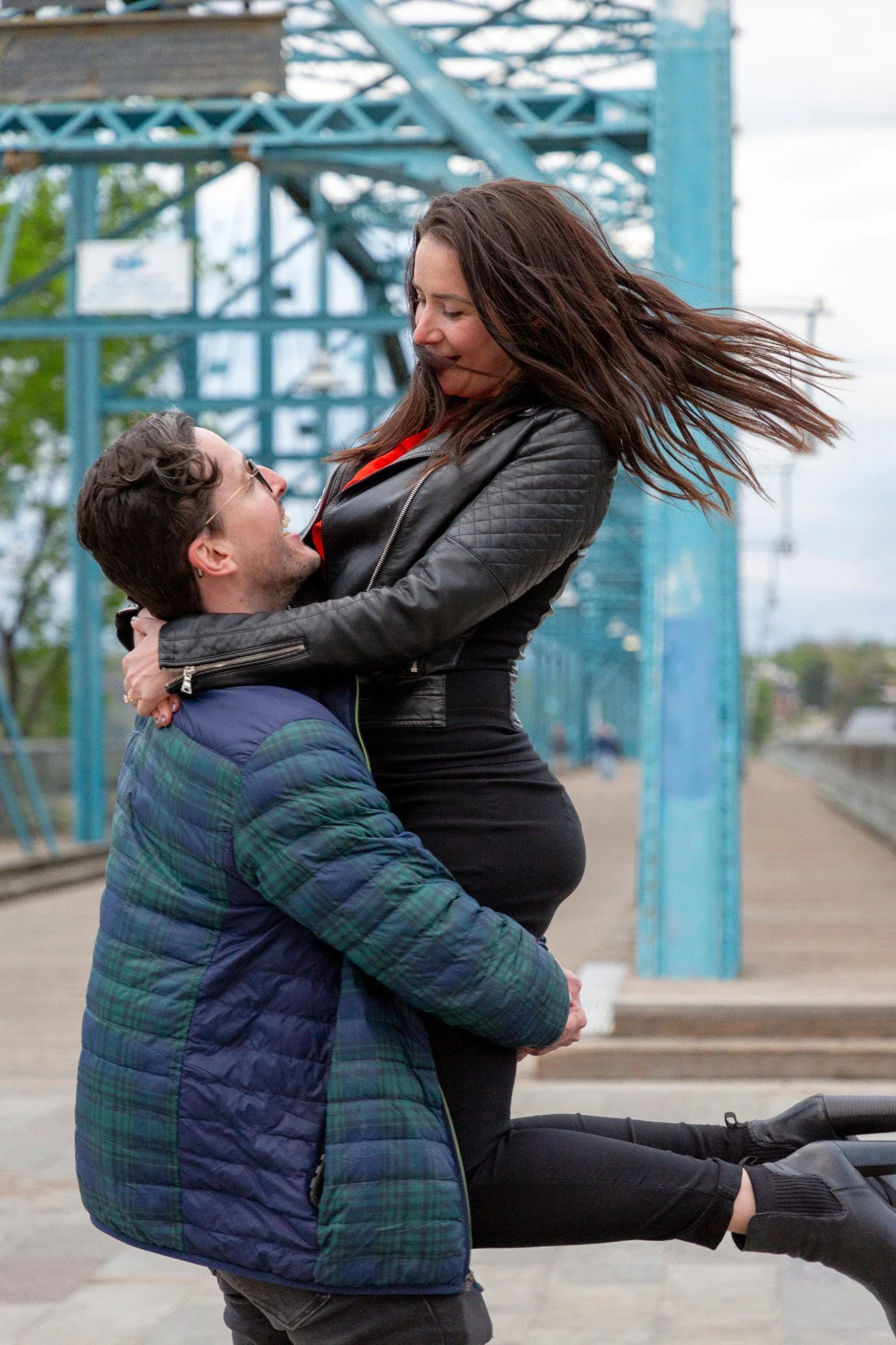 A man and a woman are outdoors. The man is kneeling and lifting the woman, who is sitting on his knees. The woman has long brown hair blowing in the wind and is wearing a black leather jacket and black pants, while the man is wearing glasses and a bl