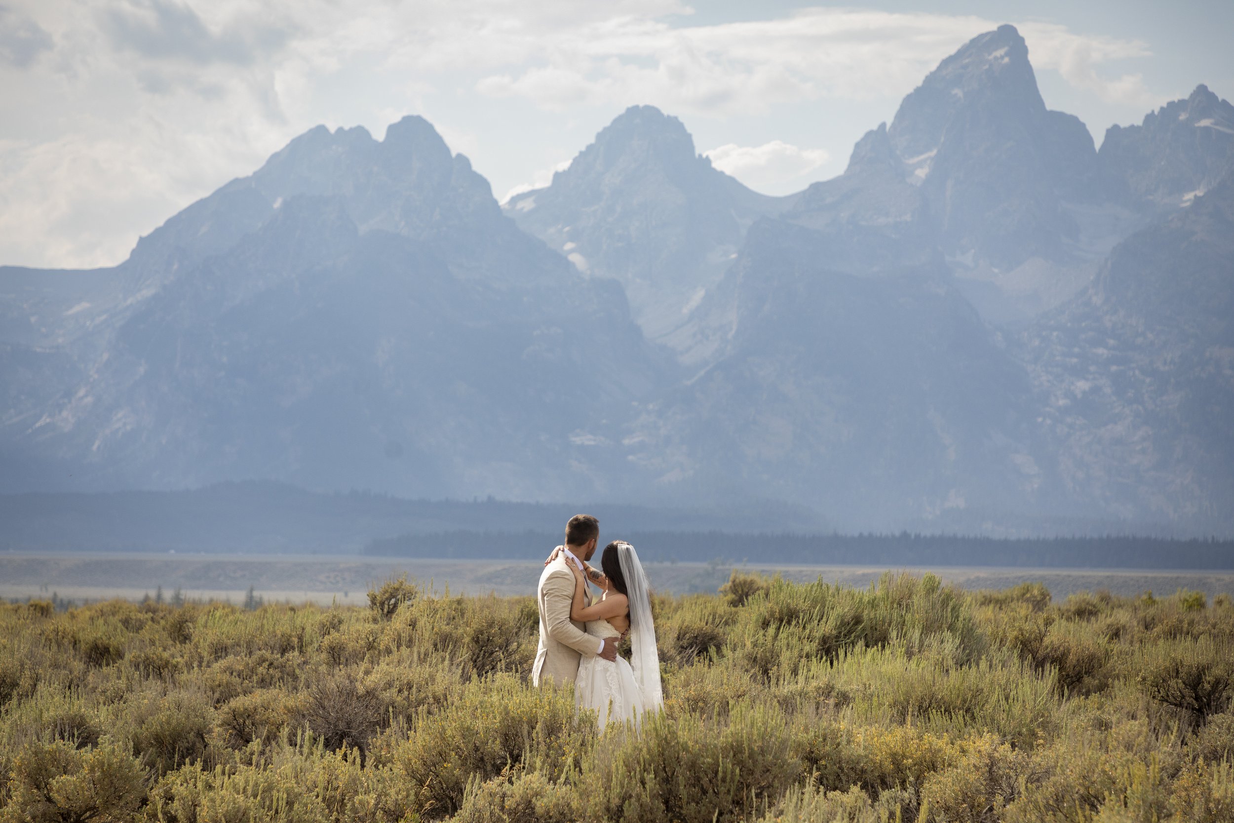 A couple in wedding attire embracing in a field with mountains in the background.