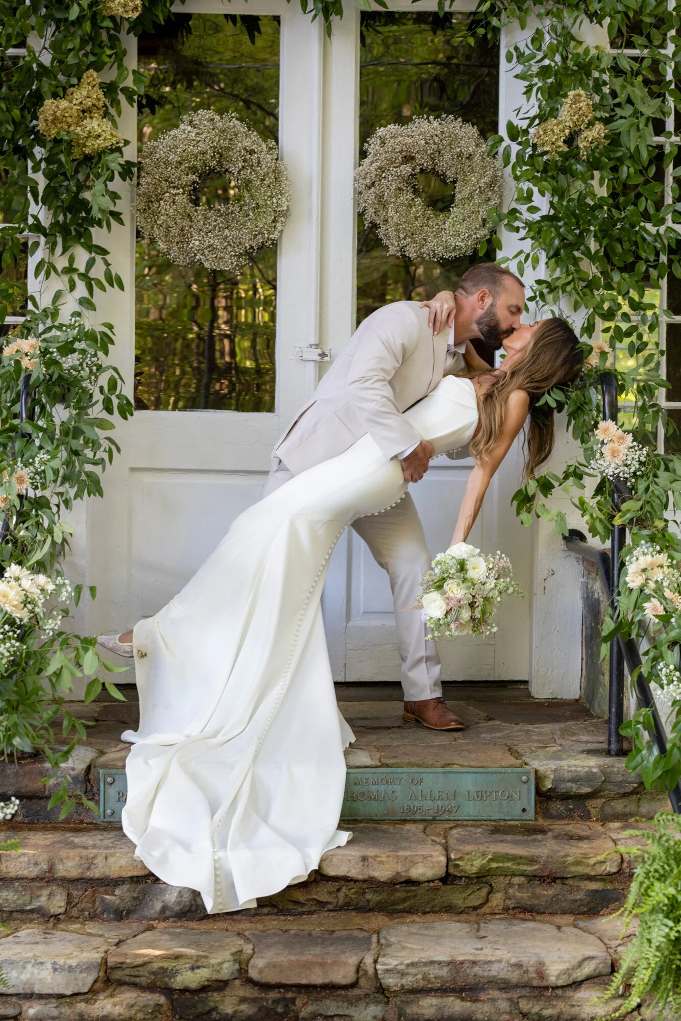A couple sharing a kiss during their wedding, standing on stone steps in front of a white door decorated with floral wreaths, surrounded by greenery.