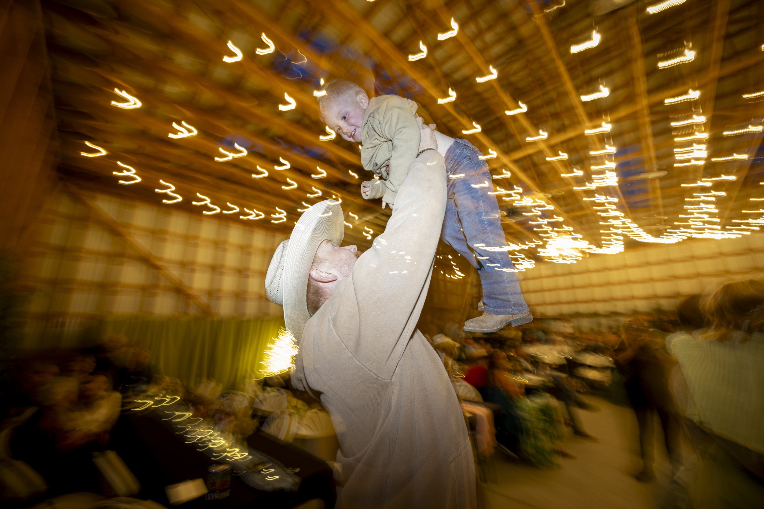 A person in a beige suit and hat lifting a smiling child in a beige jacket and jeans at an indoor carnival with bright, swirling lights overhead.