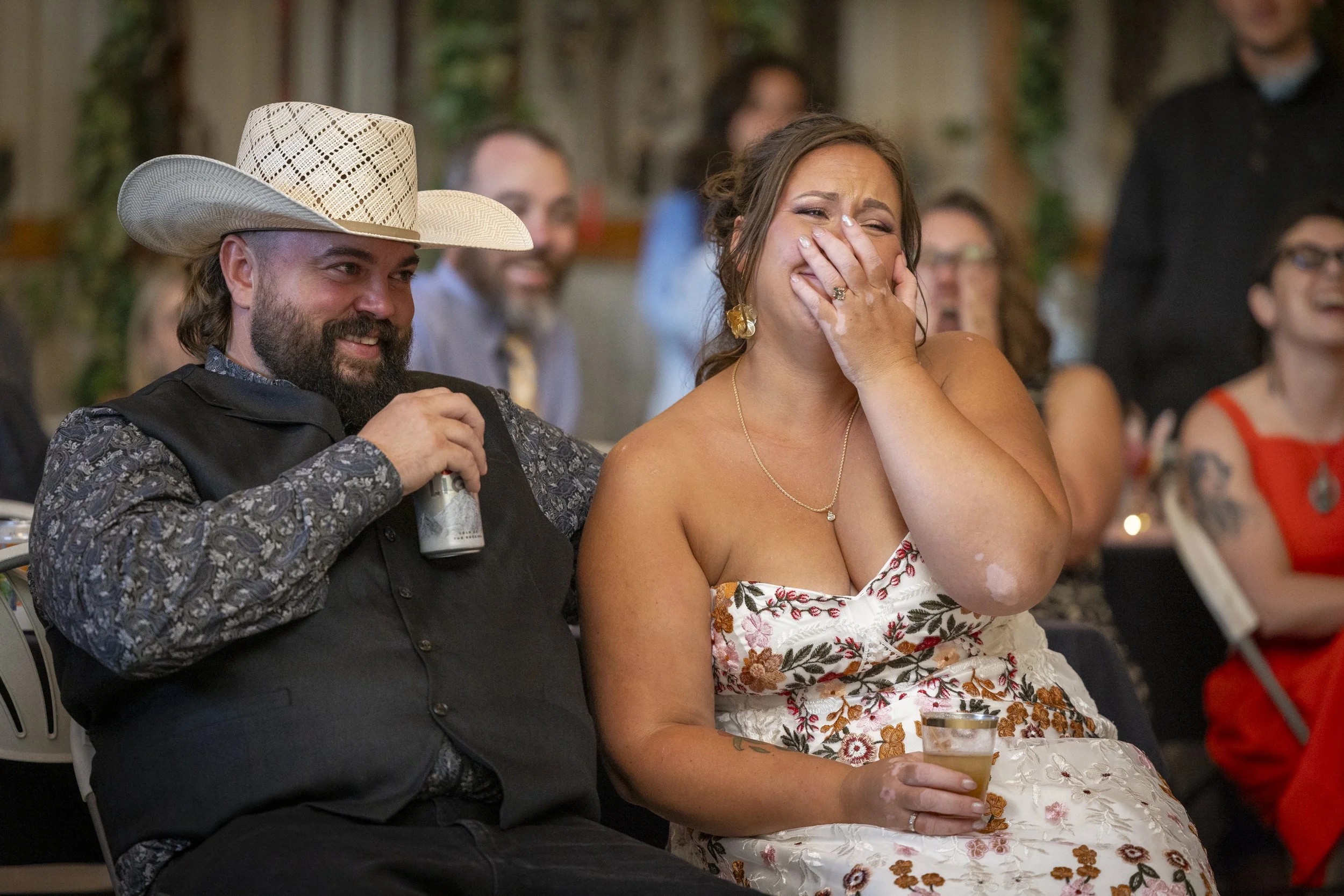 A man wearing a white cowboy hat and a floral shirt is sitting next to a woman in a strapless floral dress, who is laughing and covering her mouth with her hand. They are at a party or celebration with other people in the background.