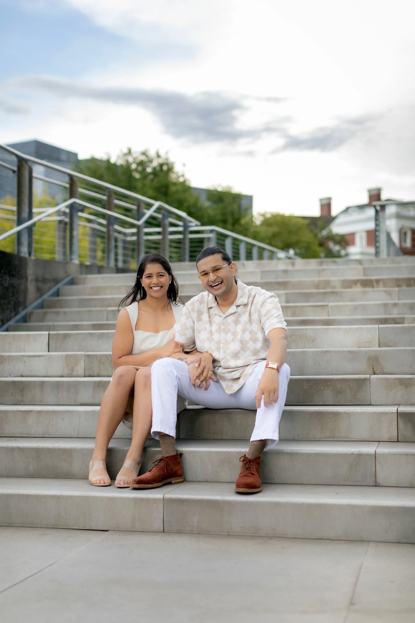 A smiling couple sitting on outdoor concrete stairs, with metal railings, in front of green trees and buildings under a cloudy sky.