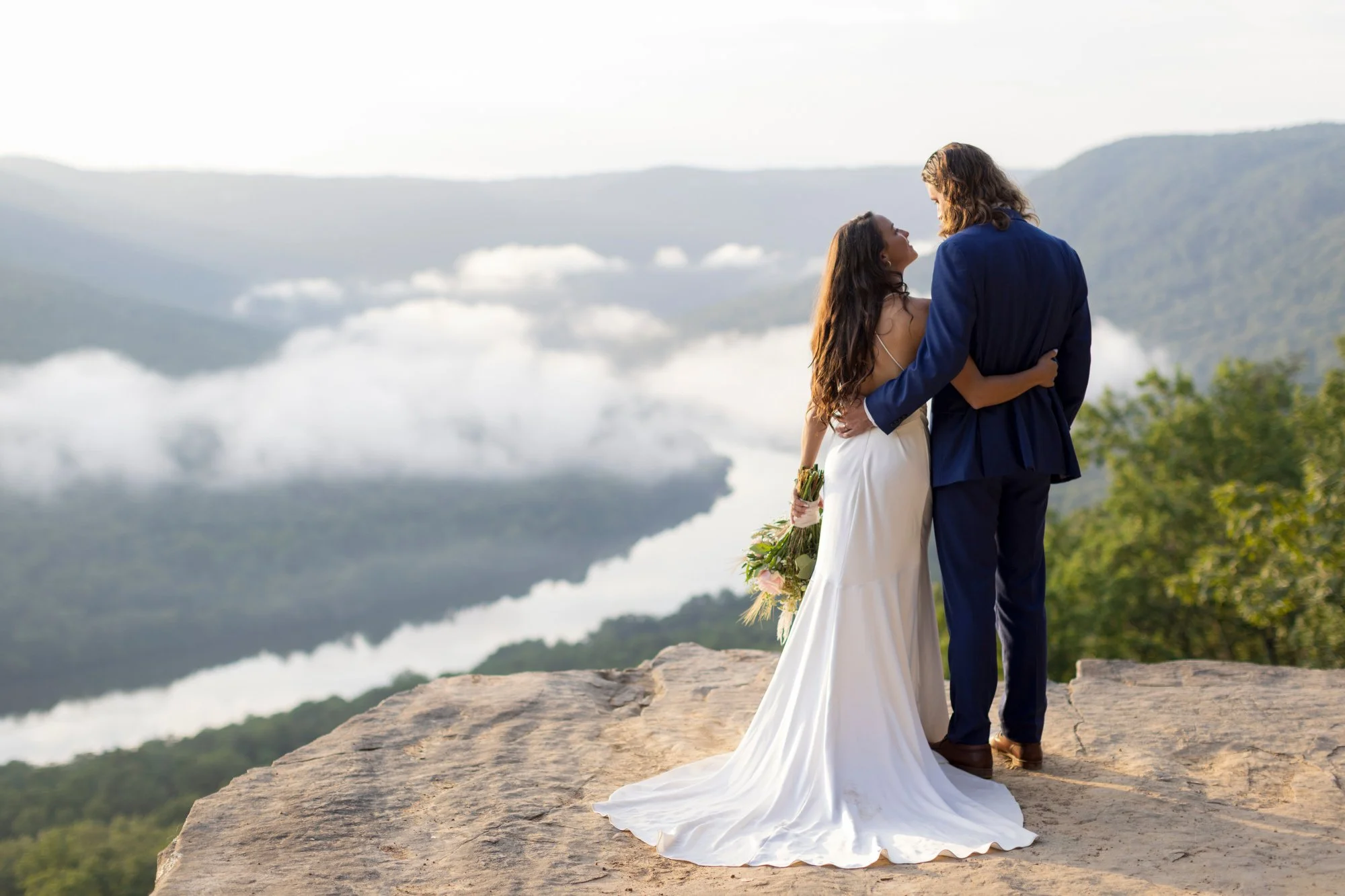 A bride and groom in wedding attire embracing each other on a cliff with mountains, clouds, and a valley in the background during daylight.