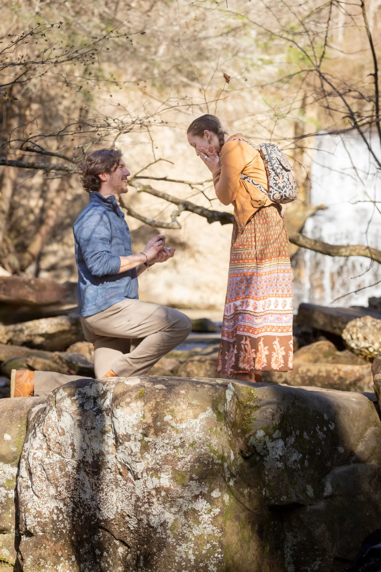 A man kneeling on a large rock proposing to a woman in a forest setting near a waterfall, with her hand covering her mouth in surprise.
