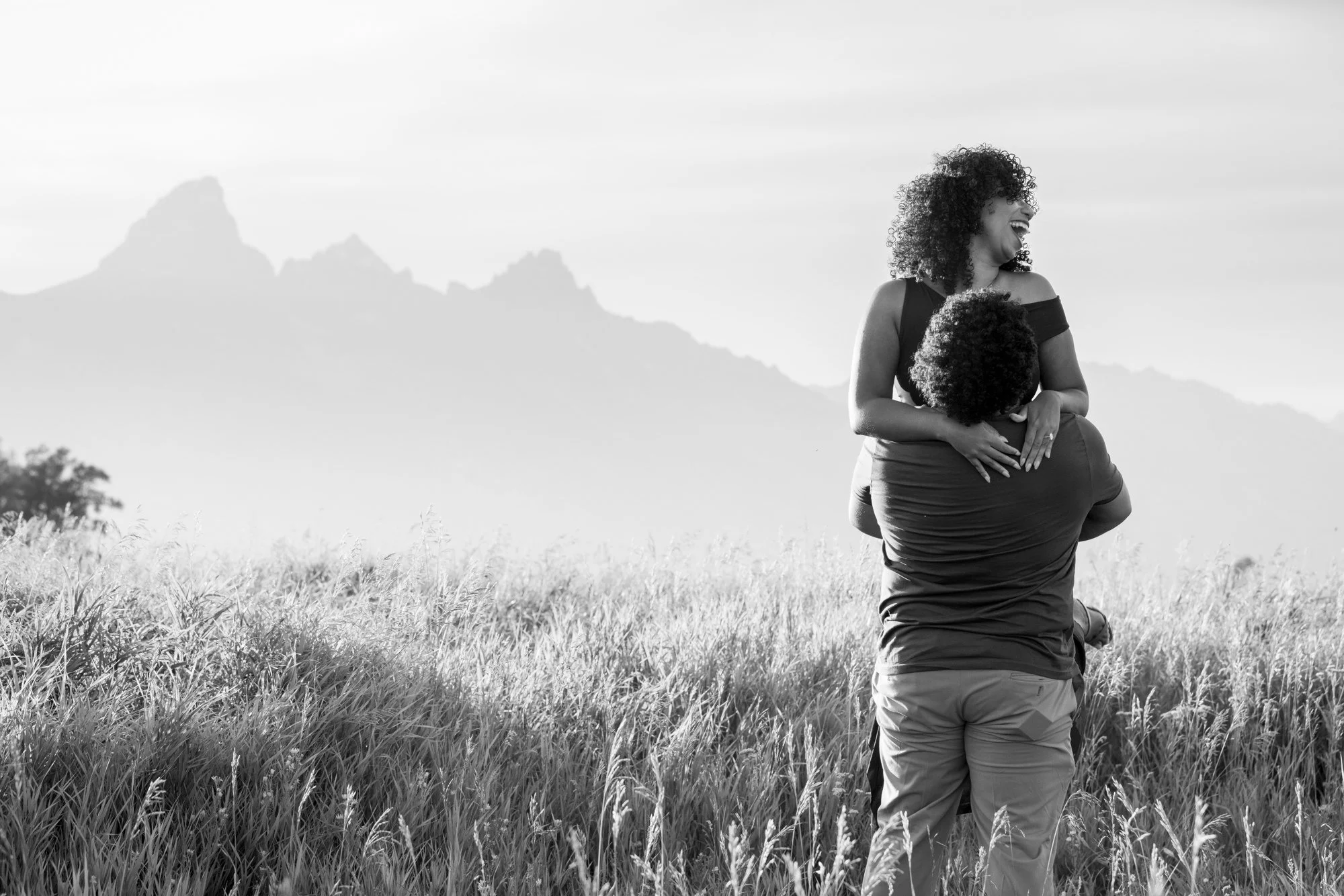 A woman with curly hair smiling and laughing while being carried on the shoulders of a man in a grassy field with mountains in the background. Black and white photo.