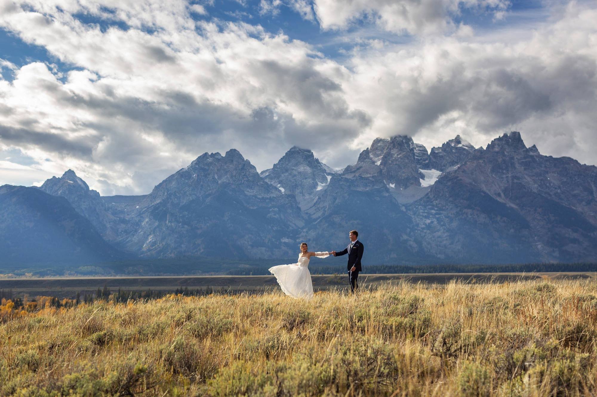 A bride and groom hold hands in a field with mountain peaks and a cloudy sky in the background.