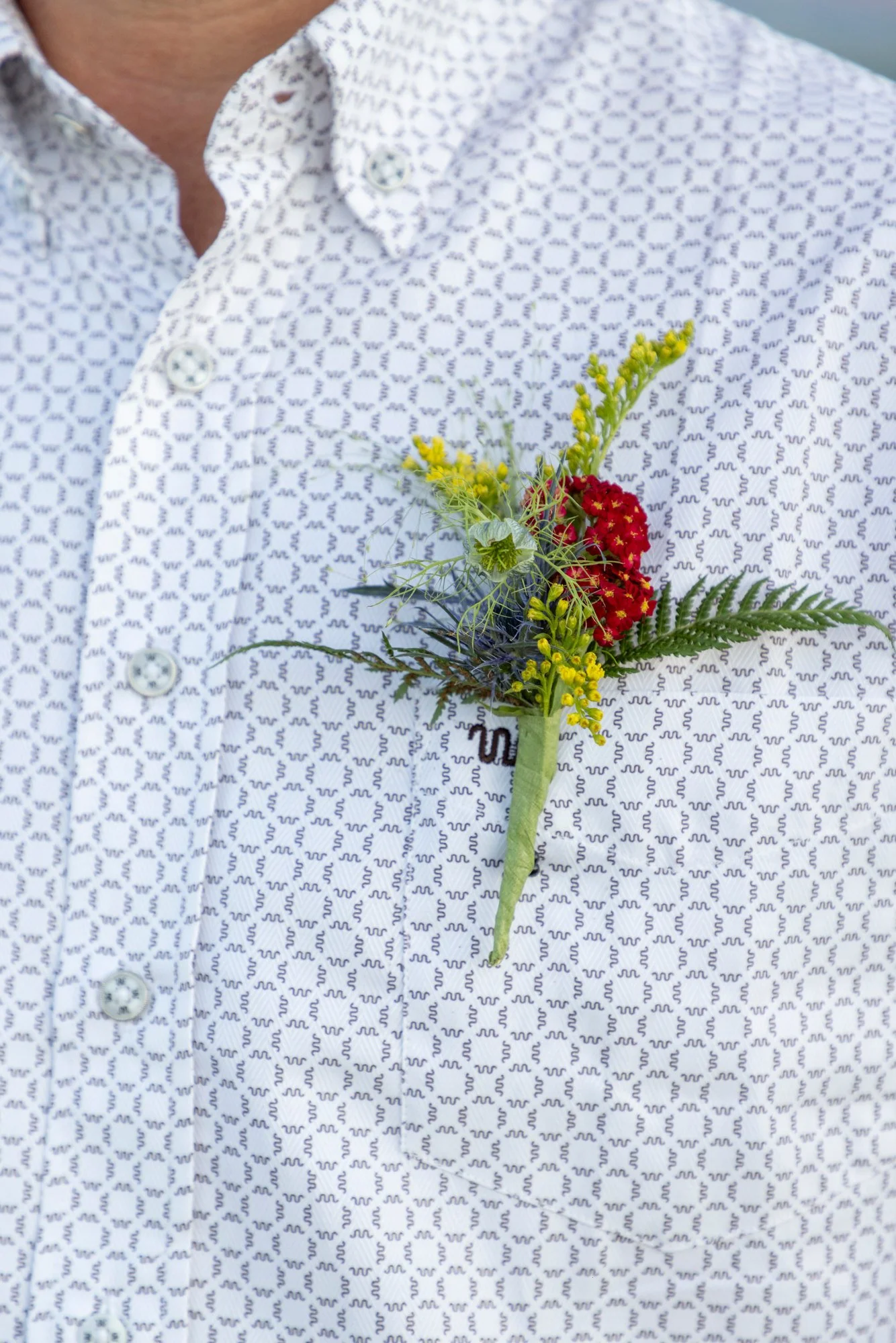A person wearing a patterned white shirt with small black squiggles and a boutonniere made of yellow and red flowers, greenery, and ferns pinned to the left side.