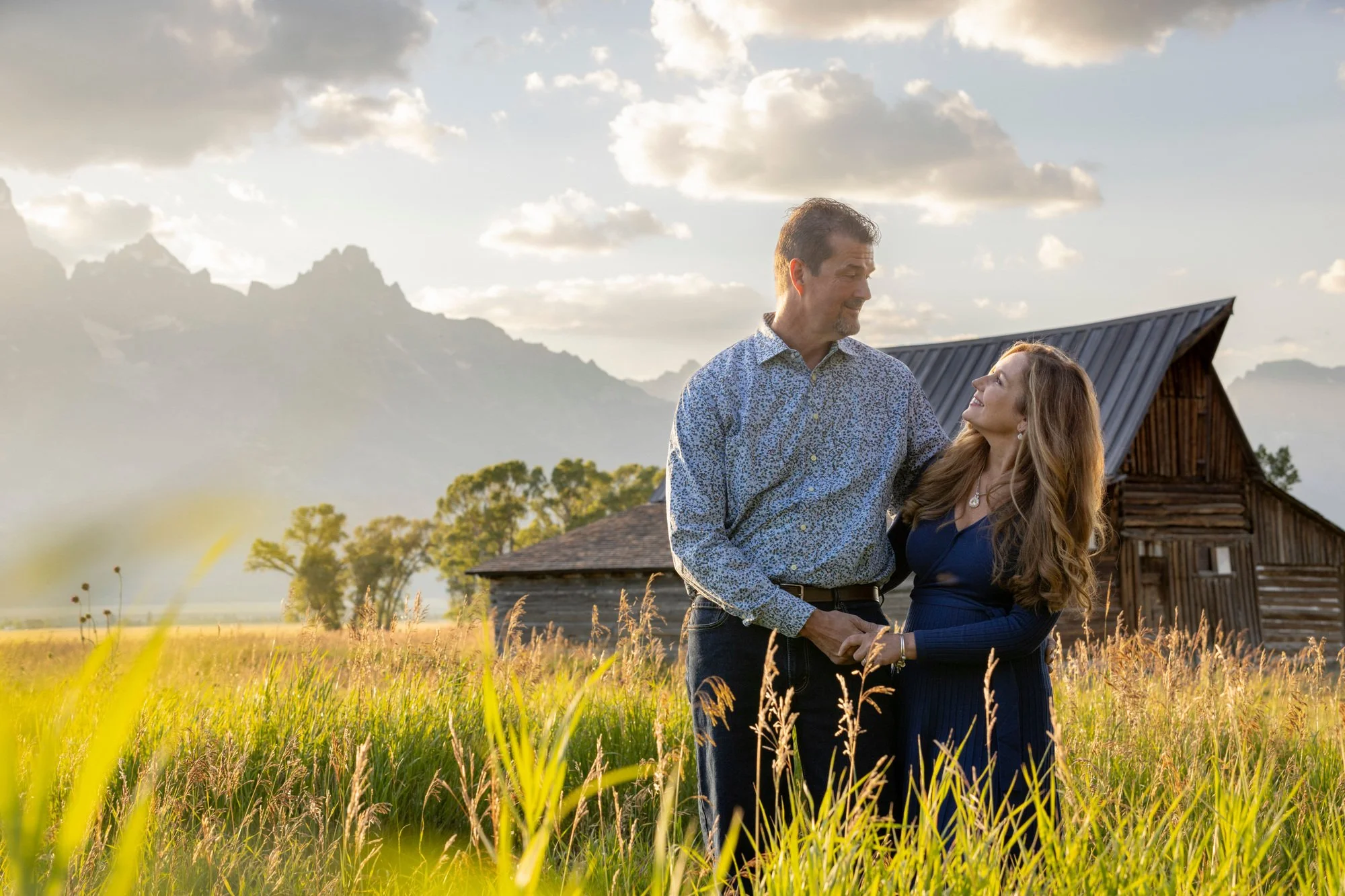 A middle-aged man and woman stand in a field of tall grass, gazing at each other and smiling. The man is wearing a patterned button-up shirt, and the woman is in a dark blue dress. In the background, there is a rustic wooden barn and mountains under 