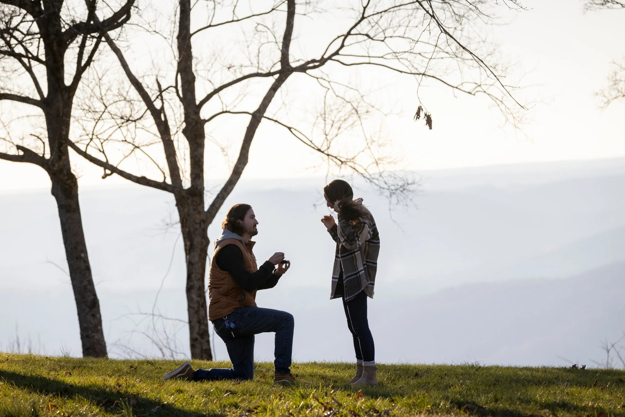 A man kneeling on one knee proposing to a woman outdoors in a grassy area with trees and snow in the background during winter, with a woman standing in front of him, holding and looking at his hand.