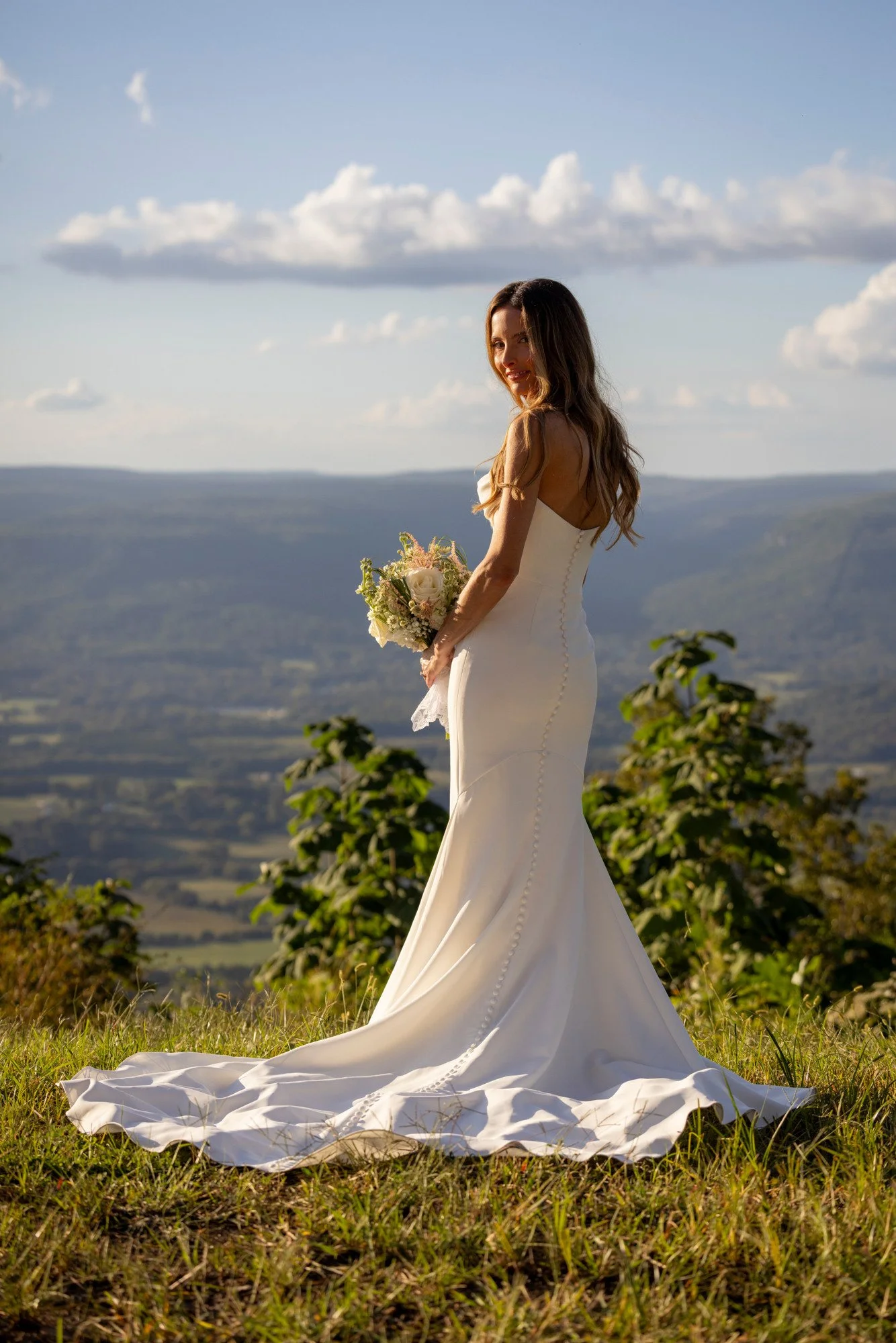 A woman in a white wedding dress holding a bouquet of flowers, standing outdoors on a grassy hillside with mountains and a partly cloudy sky in the background.