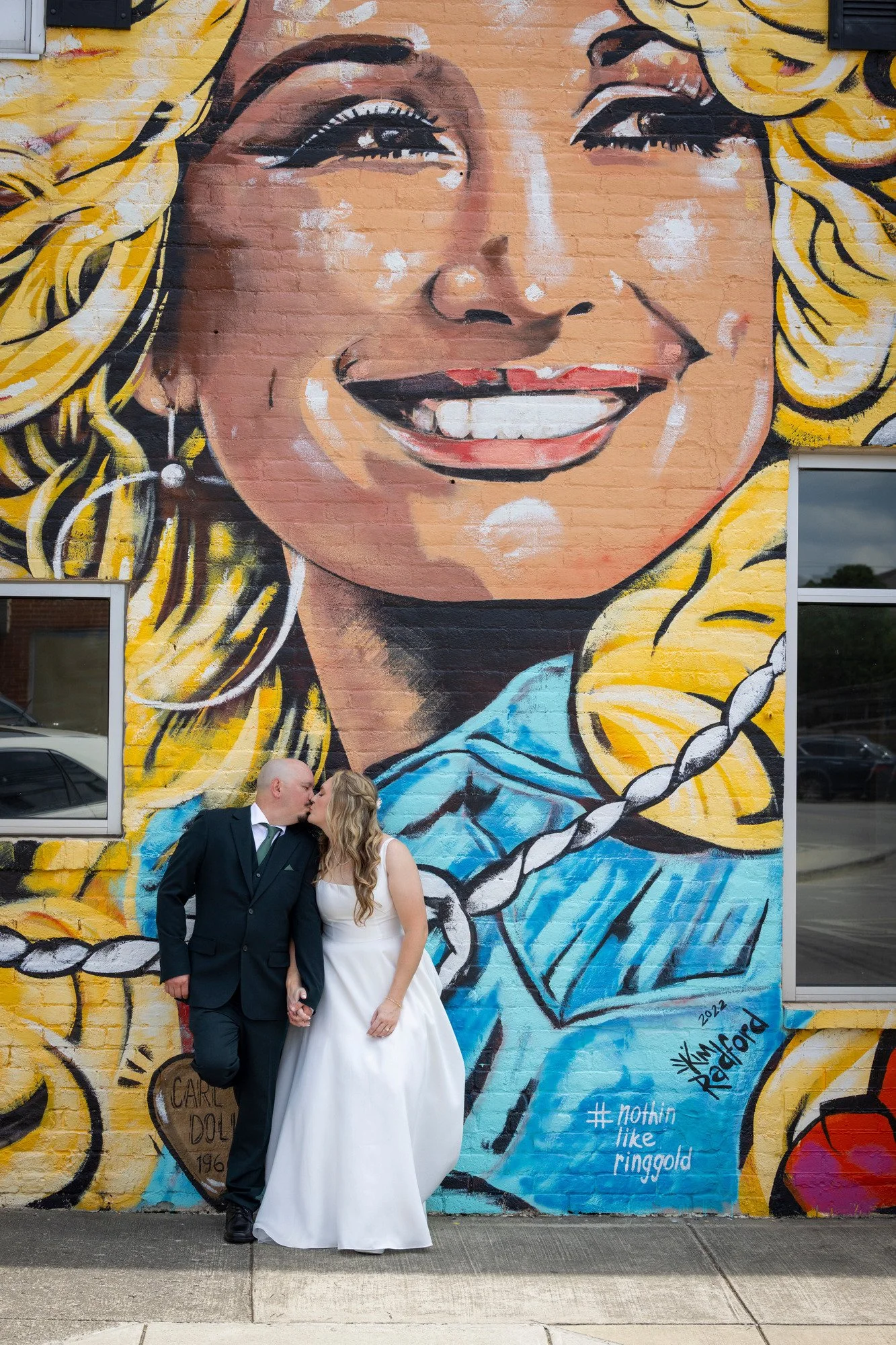 A couple in formal attire, holding hands and kissing, standing in front of a large mural of a smiling woman with blonde hair. The mural features bright colors and artistic details.