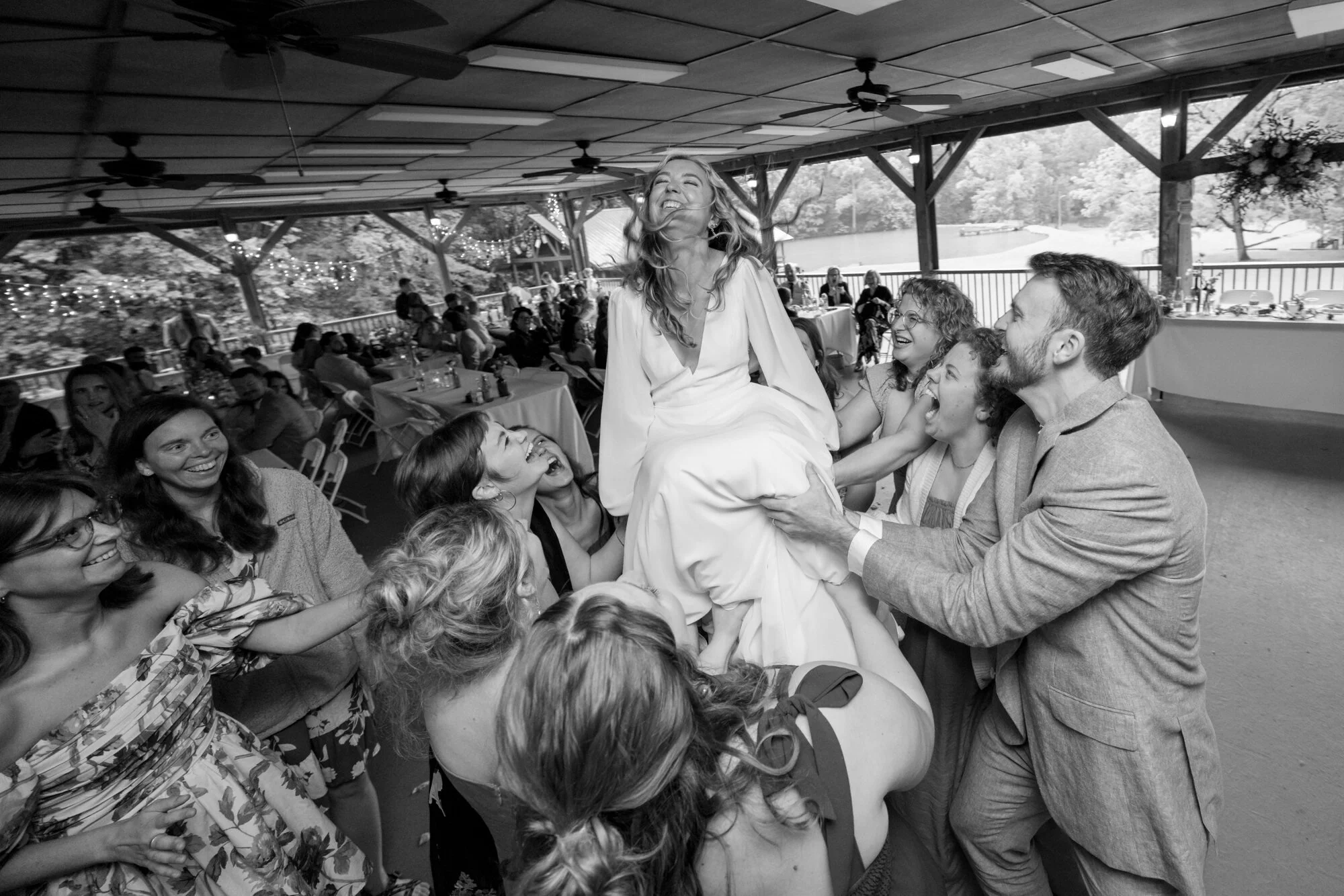 A woman in a white dress is being lifted by a group of people at an outdoor wedding reception. She appears joyful and is smiling broadly. The setting is a decorated open pavilion with tables, guests, and string lights, and trees are visible in the ba