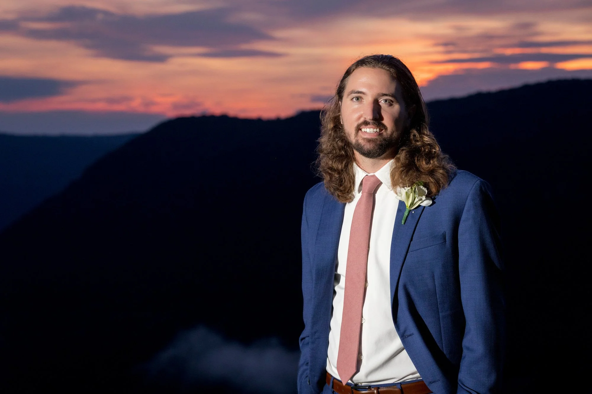 A man with long hair and a beard wearing a blue suit, white shirt, and pink tie, standing outdoors at sunset with mountains in the background.