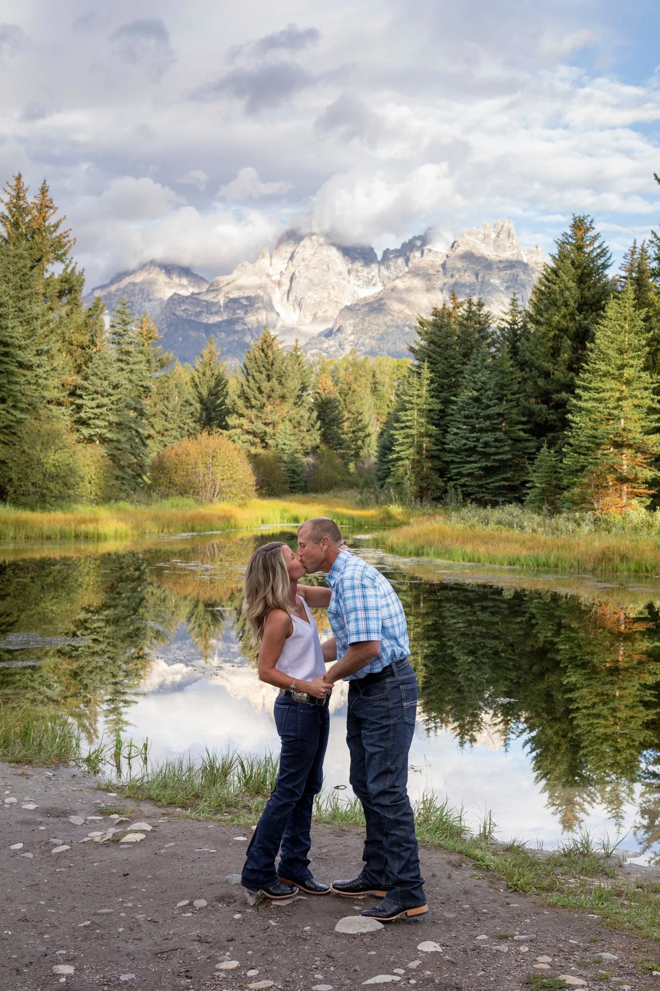 A couple kissing near a lake with a mountain range and forest in the background, under cloudy sky.