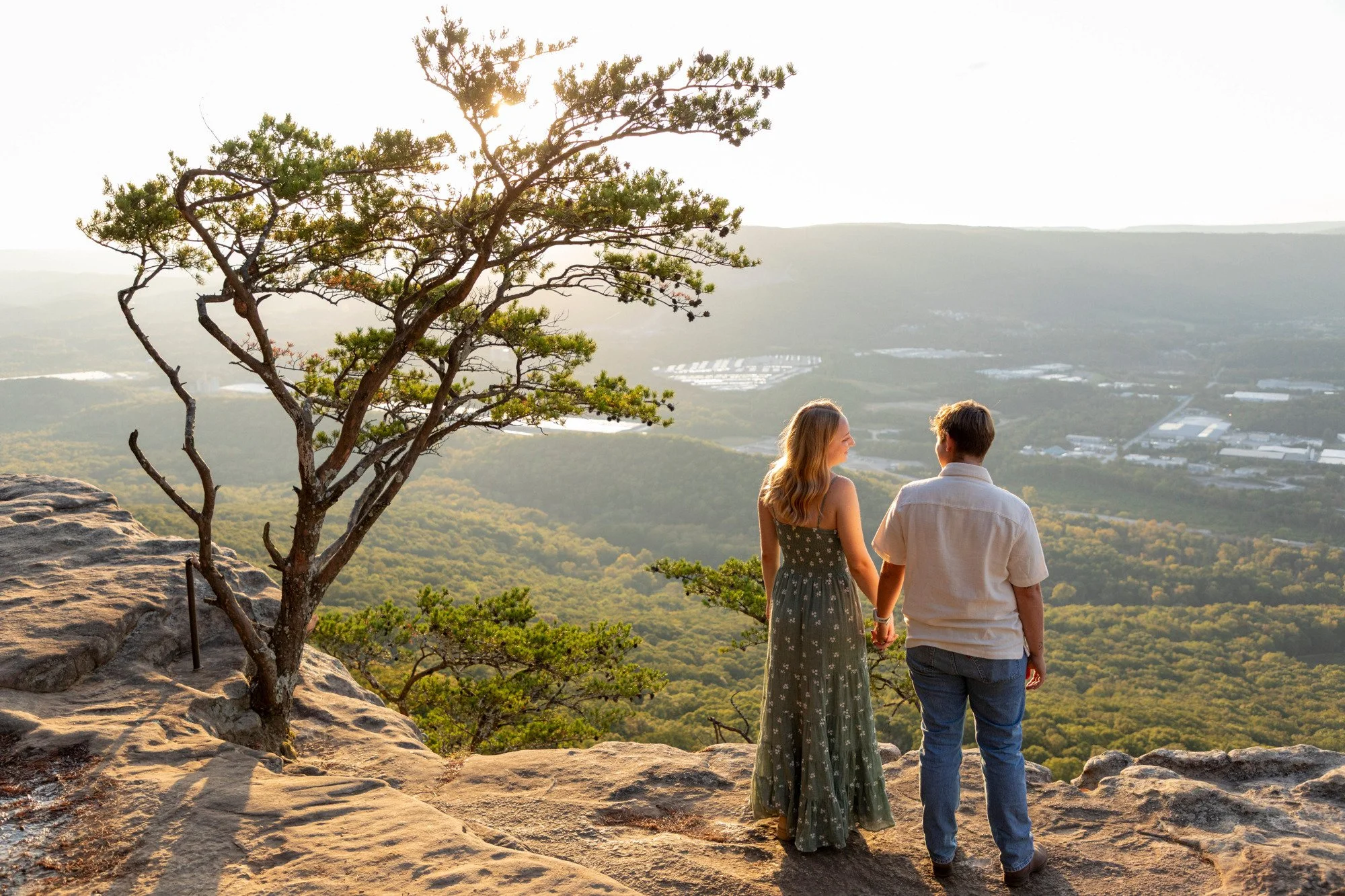 A young couple holds hands and stands on a rocky ledge overlooking a landscape at sunset, with a tree in the foreground and hills and trees in the distance.