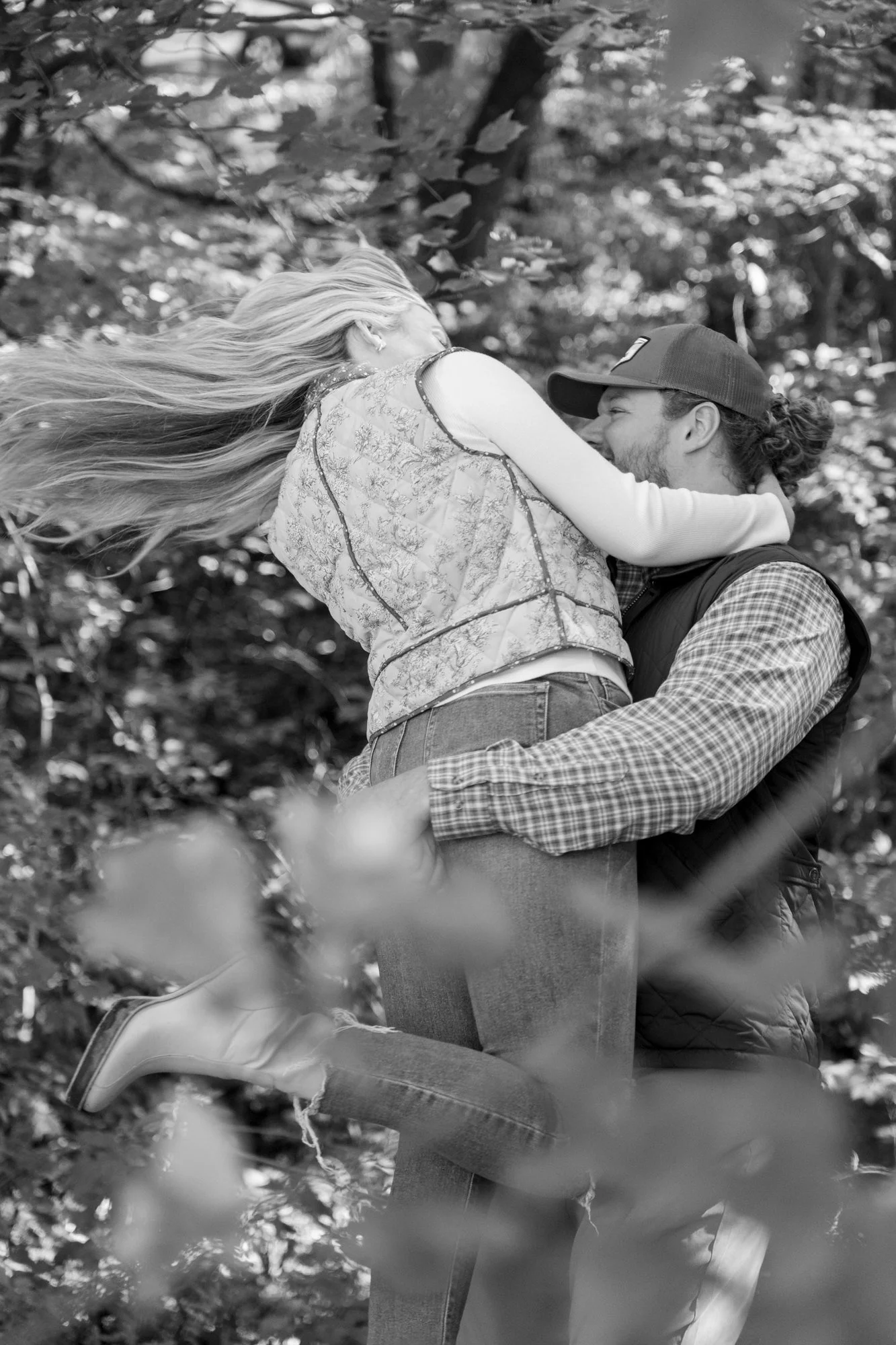 A couple in an outdoor setting, the woman with long flowing hair and the man wearing a hat, holding each other closely and smiling.