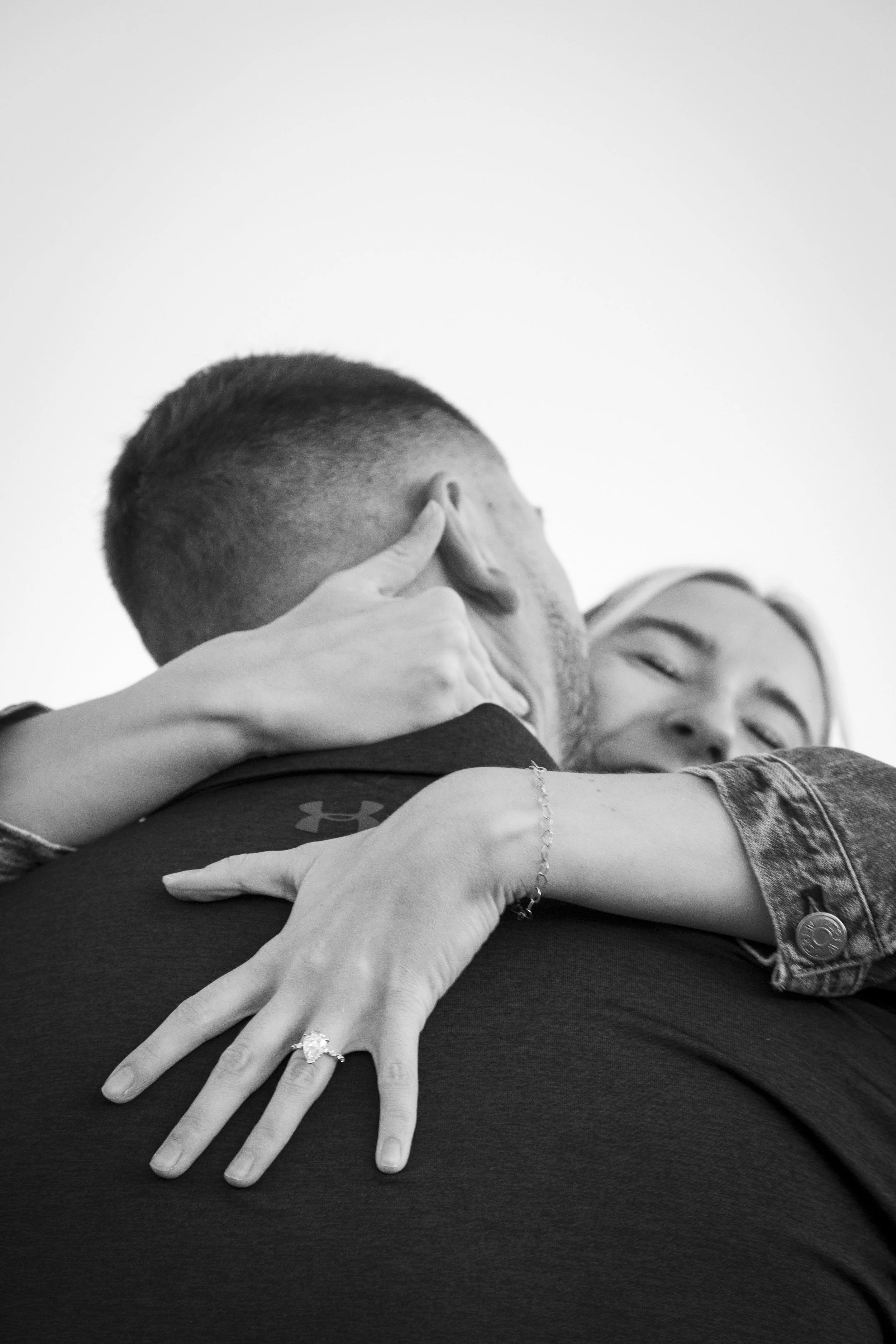 Close-up black and white photo of a couple hugging. The woman's face is visible with closed eyes, and her arm is wrapped around the man's neck. The man's head is turned away, with only part of his face visible. Both appear to be happy and content. Th