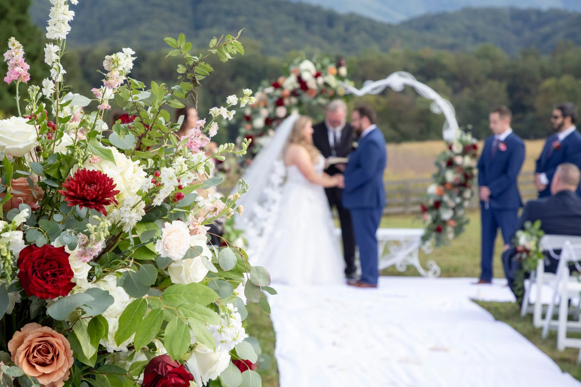 Close-up of a colorful floral arrangement with roses, peonies, and other flowers at an outdoor wedding ceremony. In the background, the officiant and couple exchange vows under a floral arch, with groomsmen and guests seated nearby in a scenic mounta