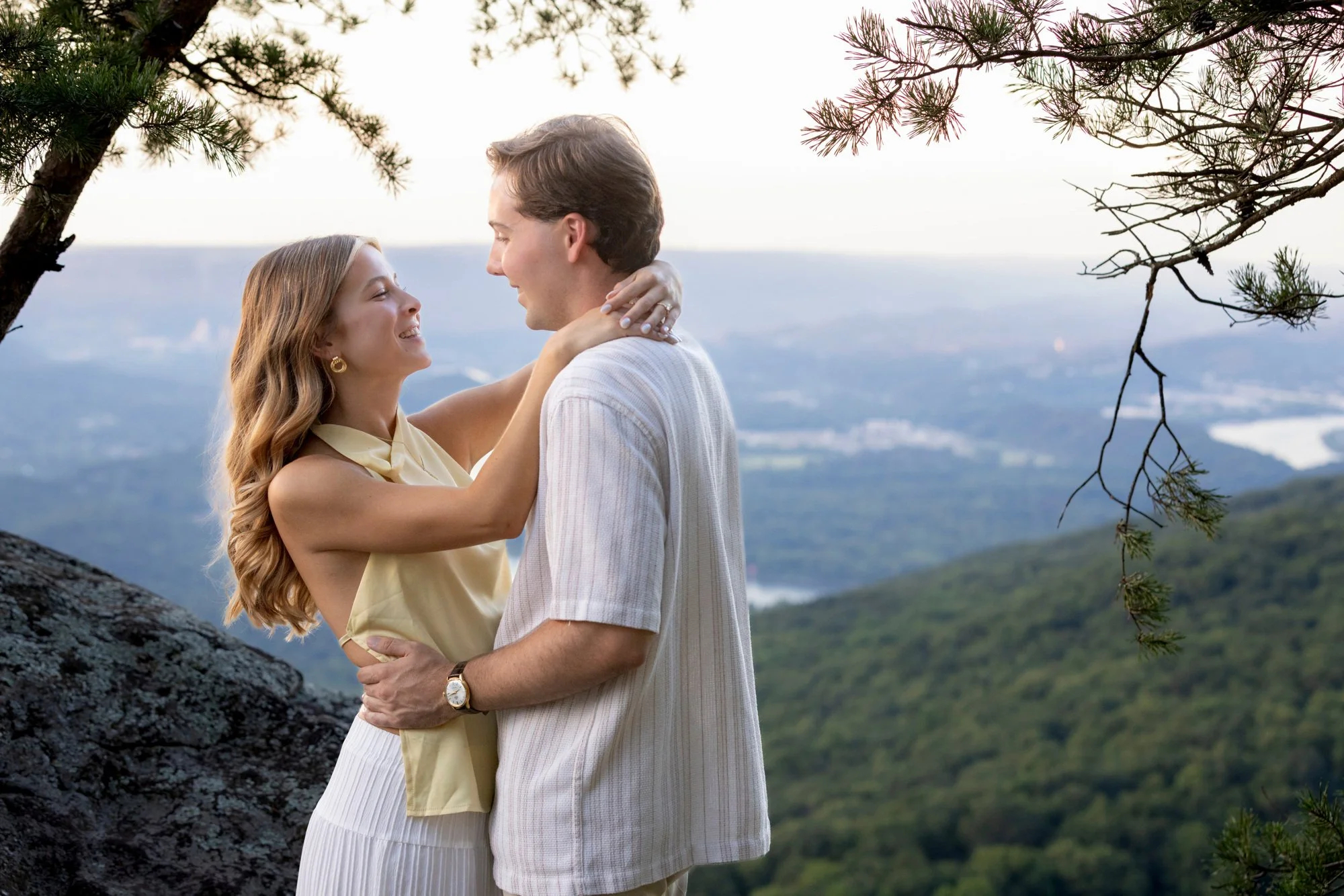 A couple embracing outdoors during daytime, surrounded by trees and overlooking a scenic background with mountains and a distant body of water.
