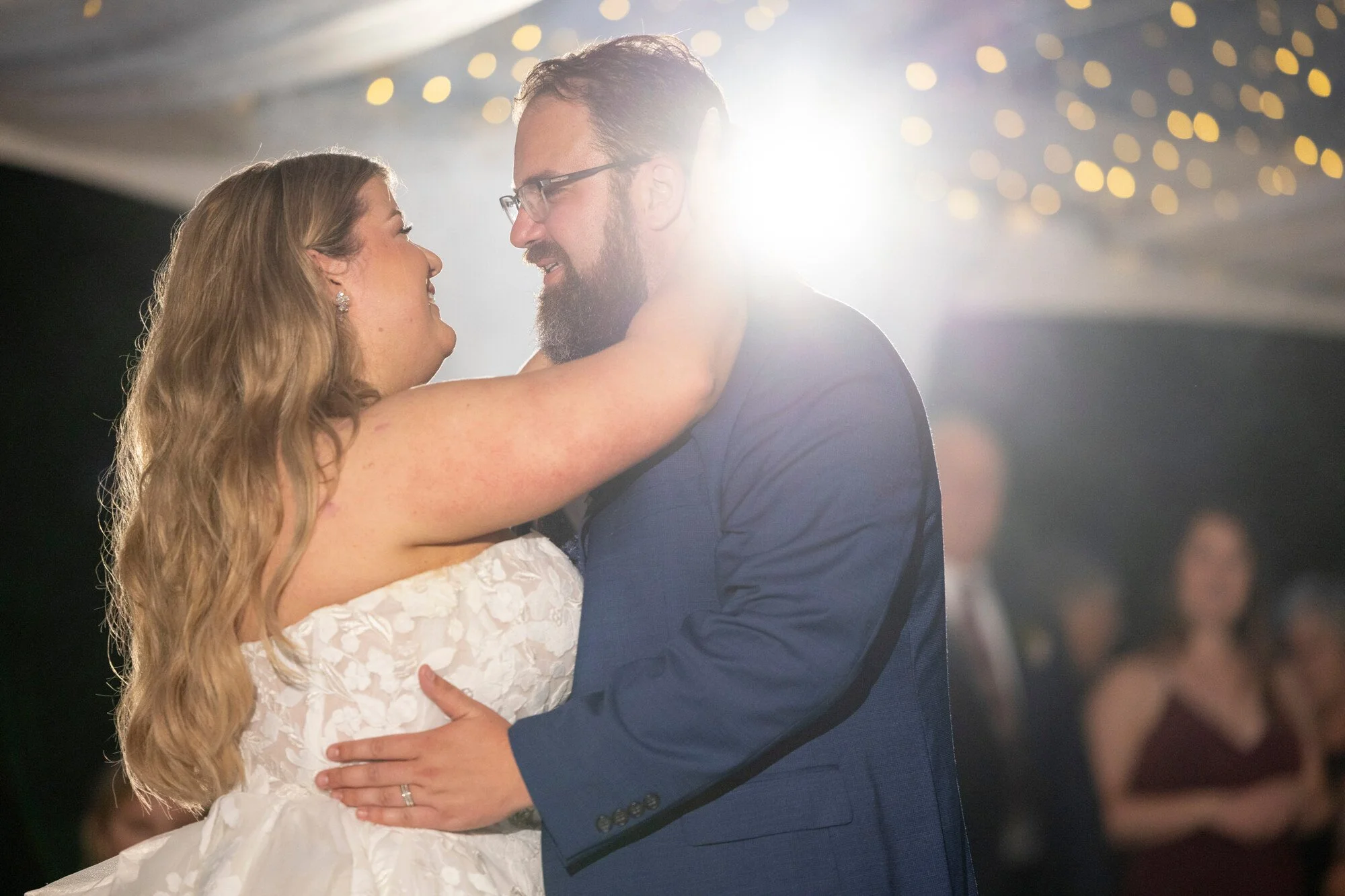 A bride and groom sharing a dance at their wedding reception, with the bride in a white lace wedding dress and the groom in a navy blue suit, smiling and looking at each other amid festive lights.