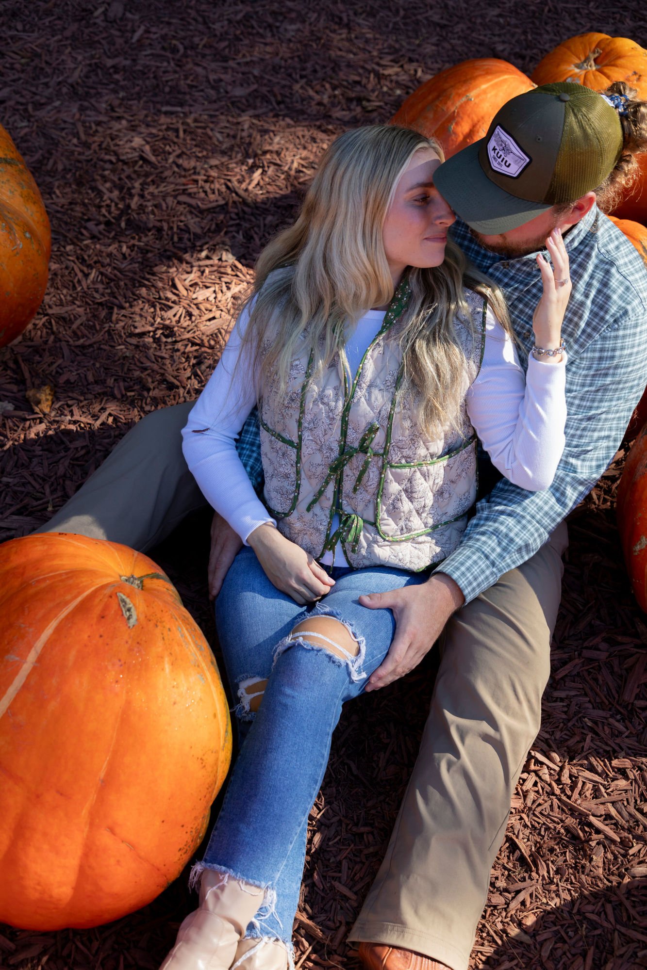 A couple sitting on the ground surrounded by pumpkins, with their foreheads touching, appearing intimate and affectionate.