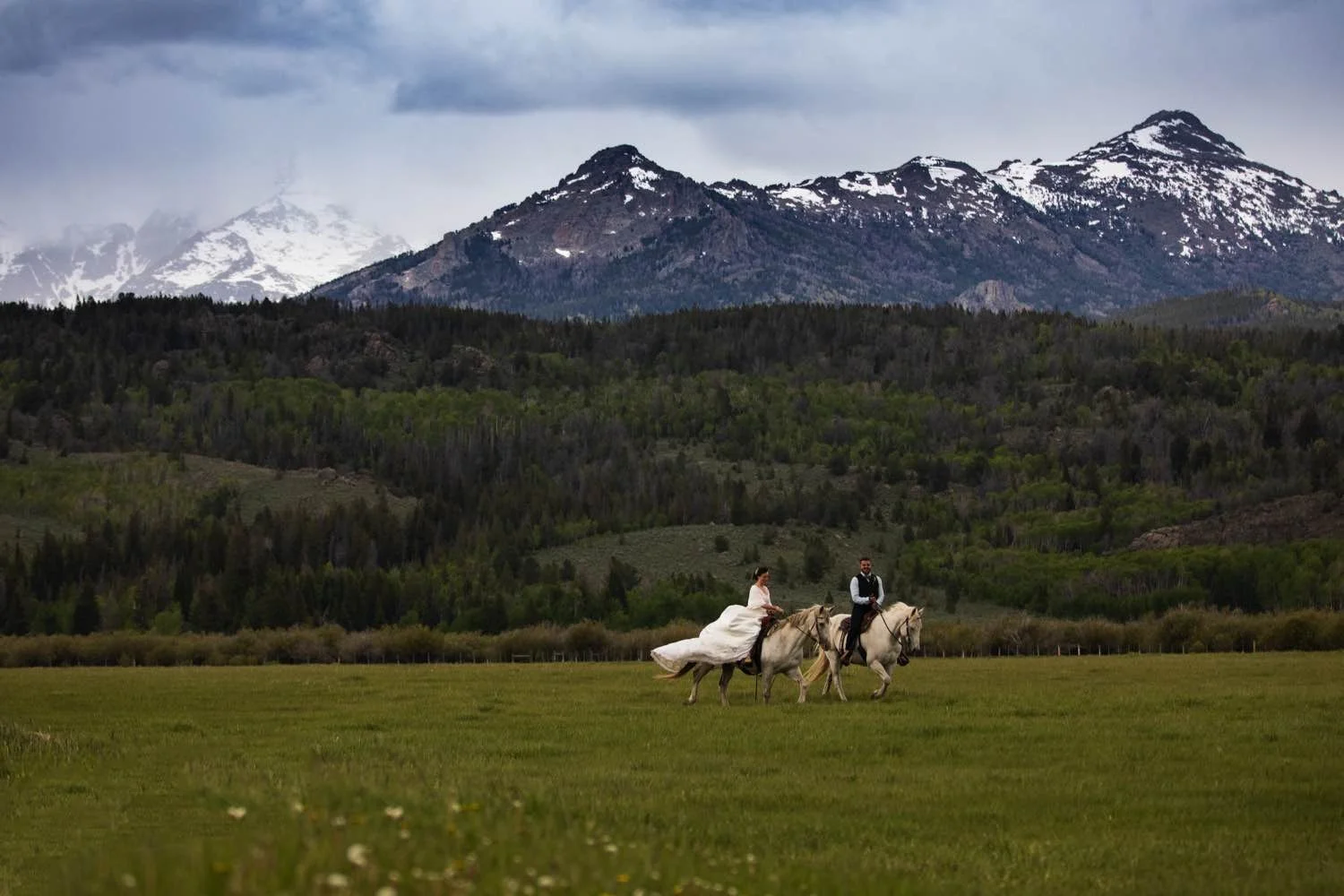 A bride and groom riding horses across a green field with mountains in the background.
