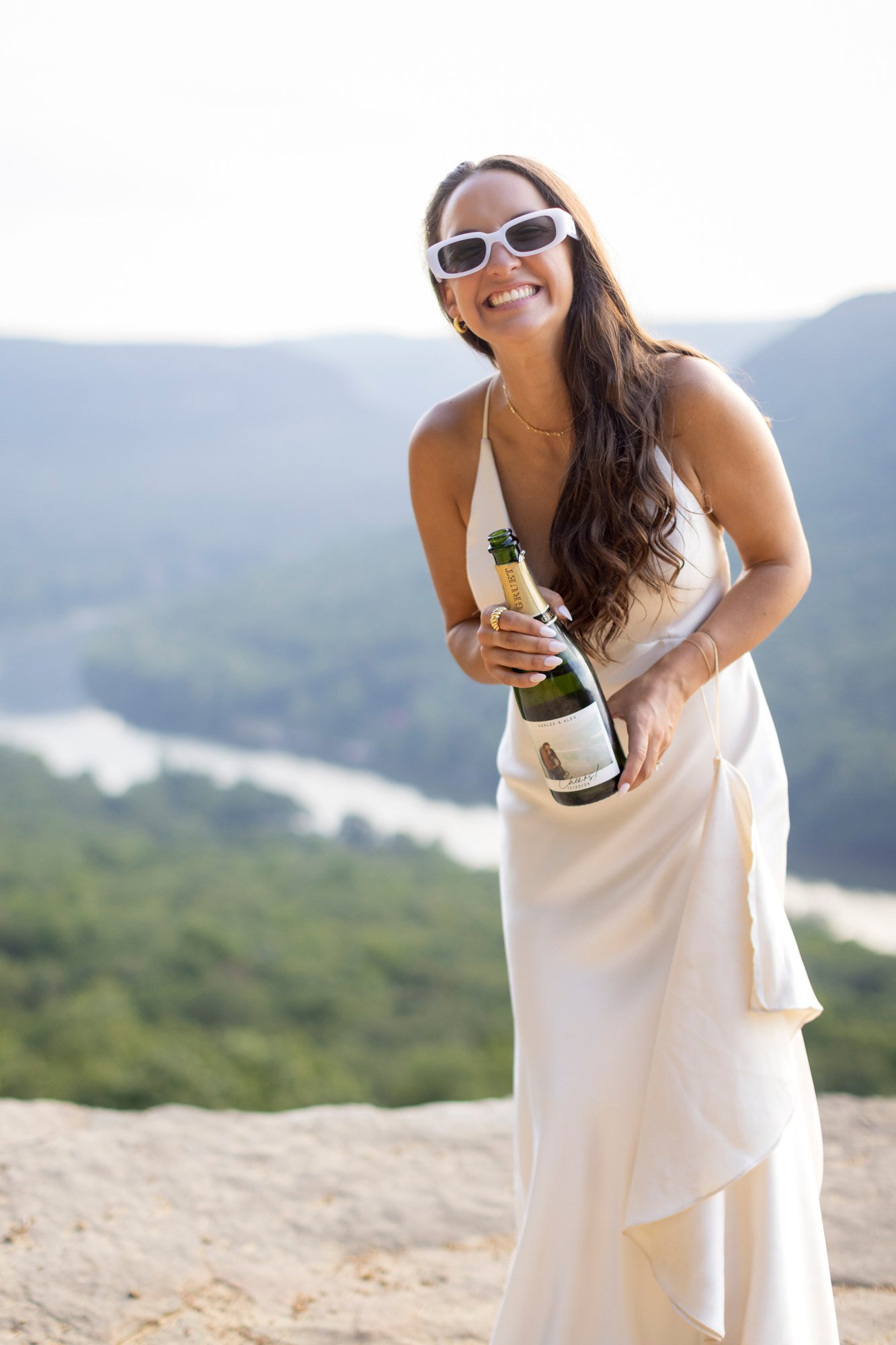 A woman in a cream dress wearing sunglasses, smiling and holding a bottle of champagne outside with mountains and a cloudy sky in the background.