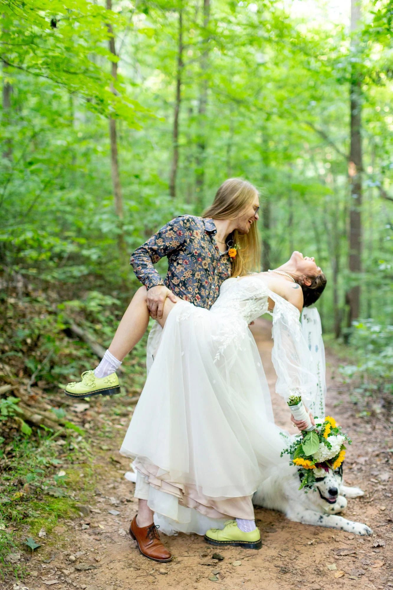 Two people, one in a wedding dress and one in casual clothing, are outdoors on a wooded path with green trees. The one in the wedding dress is lying on the ground holding a bouquet, while the other person is smiling and lifting her. A large dog with 