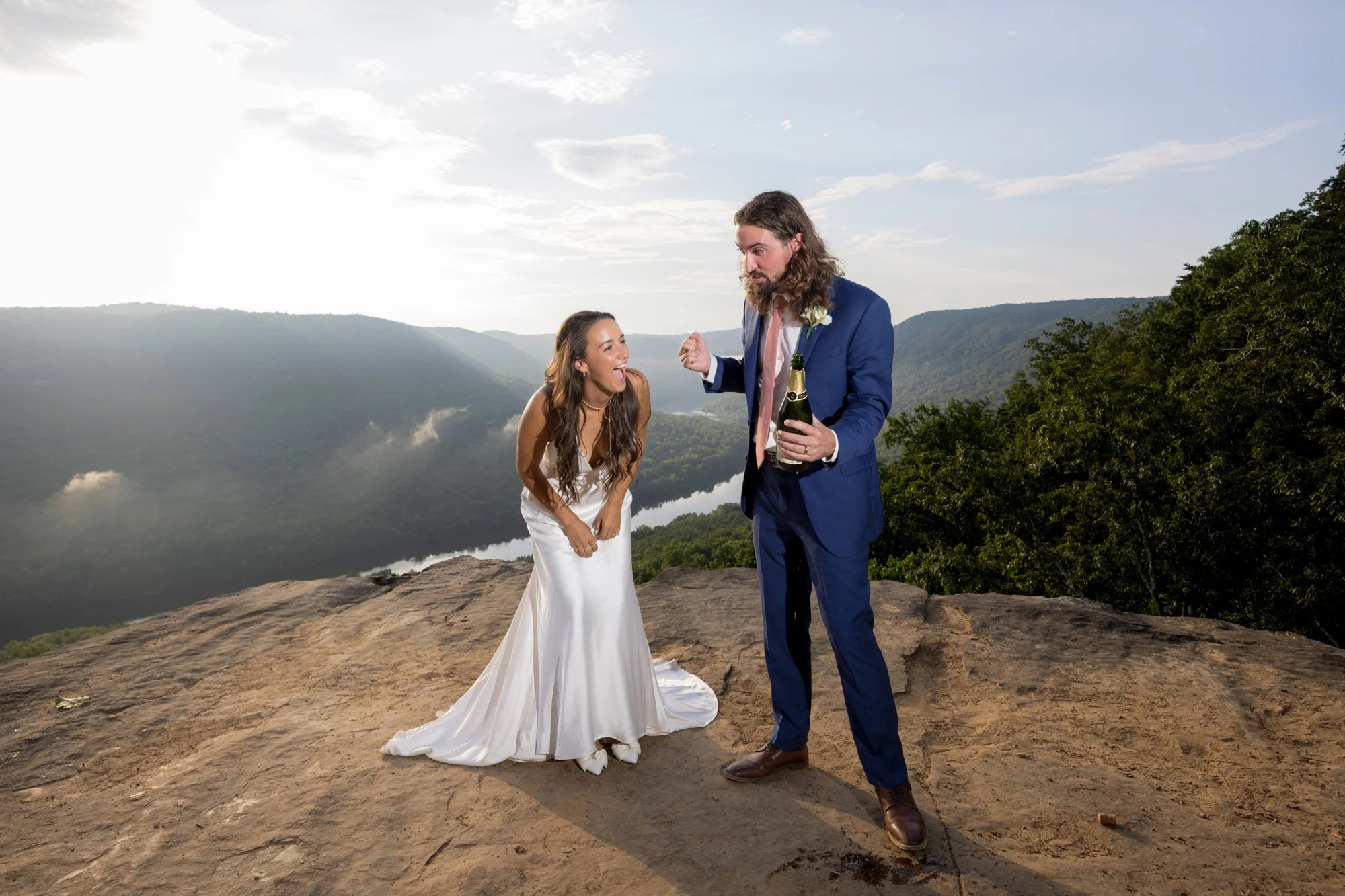A couple dressed in wedding attire celebrating outdoors on a rocky cliff with a scenic mountain and river view in the background. The woman is wearing a white wedding dress, and the man is in a blue suit holding a champagne bottle.