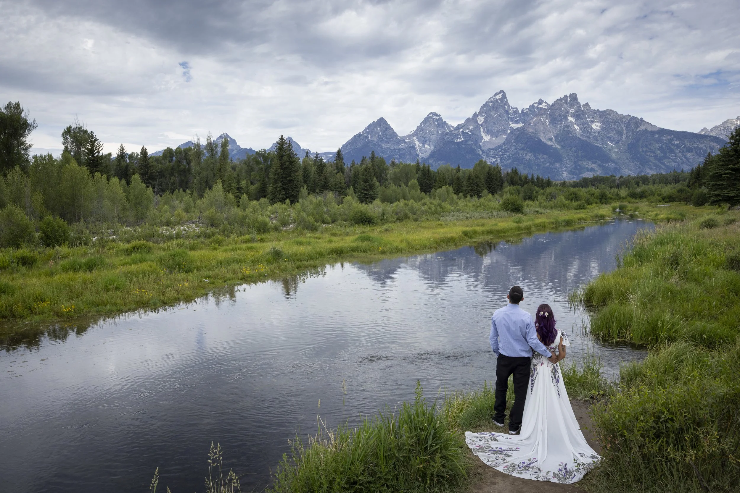 A couple dressed in wedding attire holding hands by a river with mountains and trees in the background.
