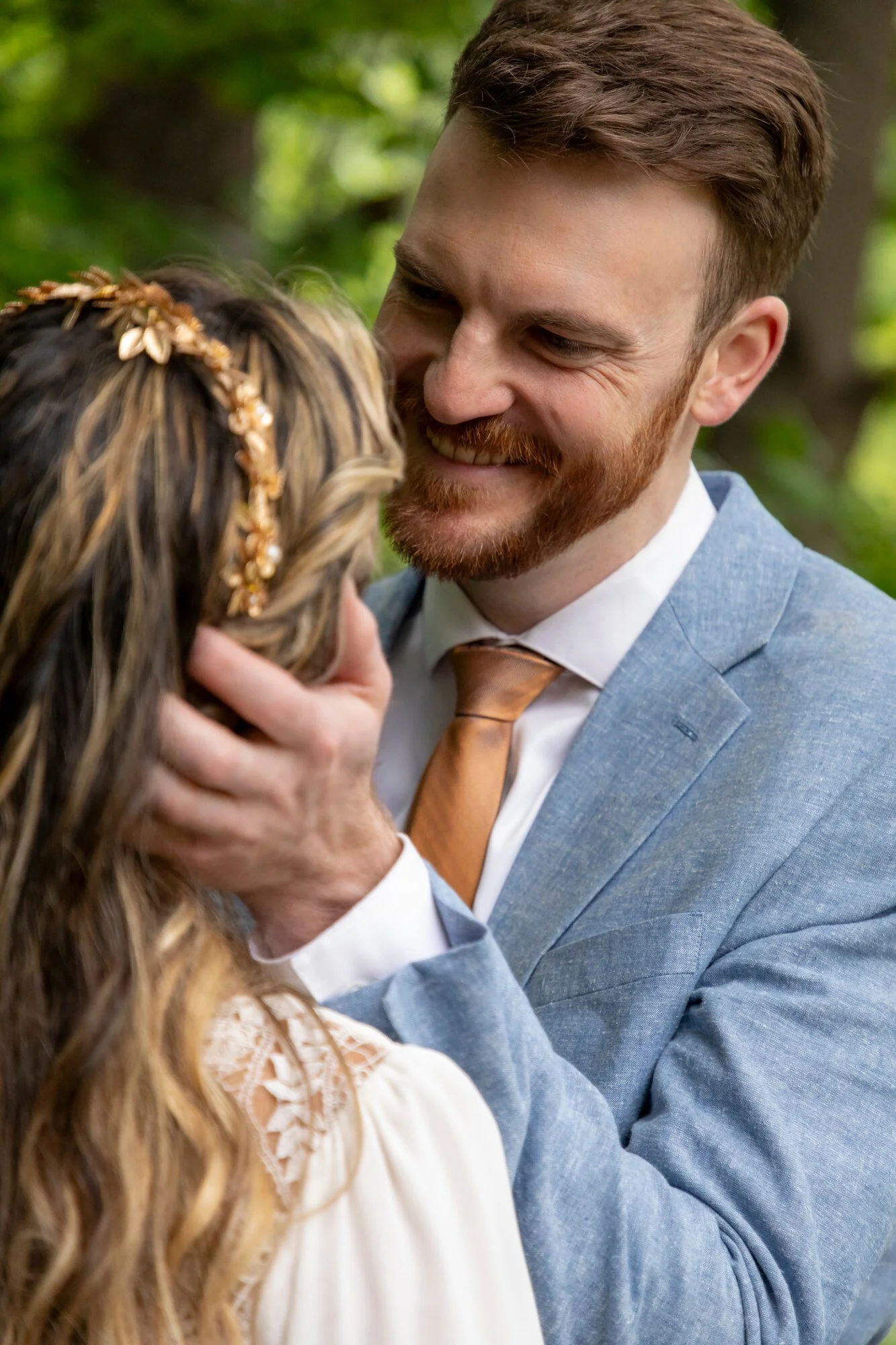 A man with brown hair and a beard, wearing a blue suit and orange tie, smiling while gently holding a woman's face in an outdoor setting with green trees in the background.