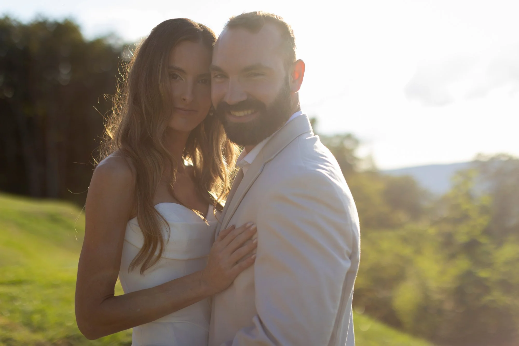 A couple dressed in wedding attire embracing outdoors during daytime, with sunlight shining behind them and greenery in the background.