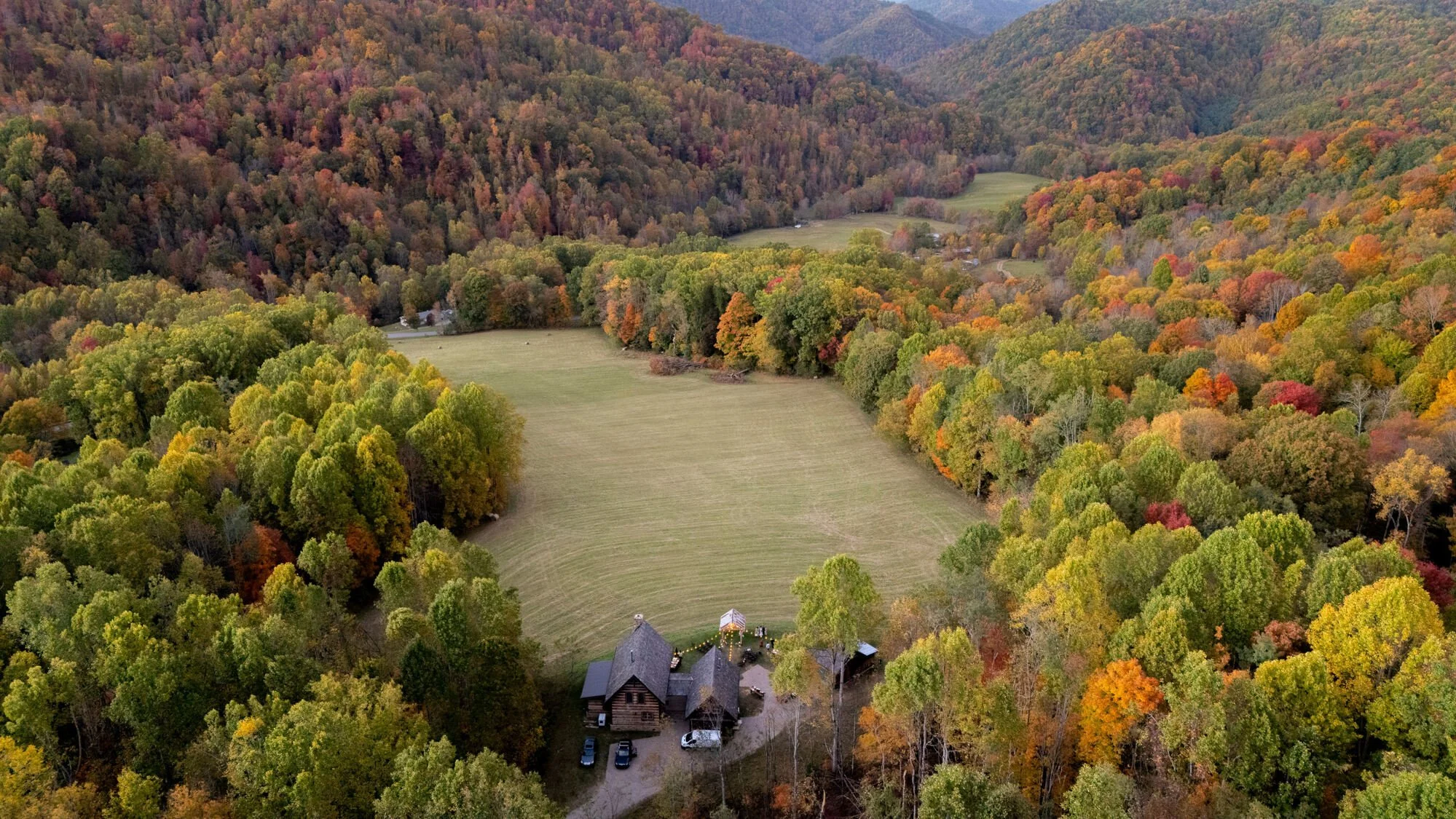 Aerial view of a mountainous landscape with colorful autumn trees, a cleared green field, and a small rustic building near the foreground.