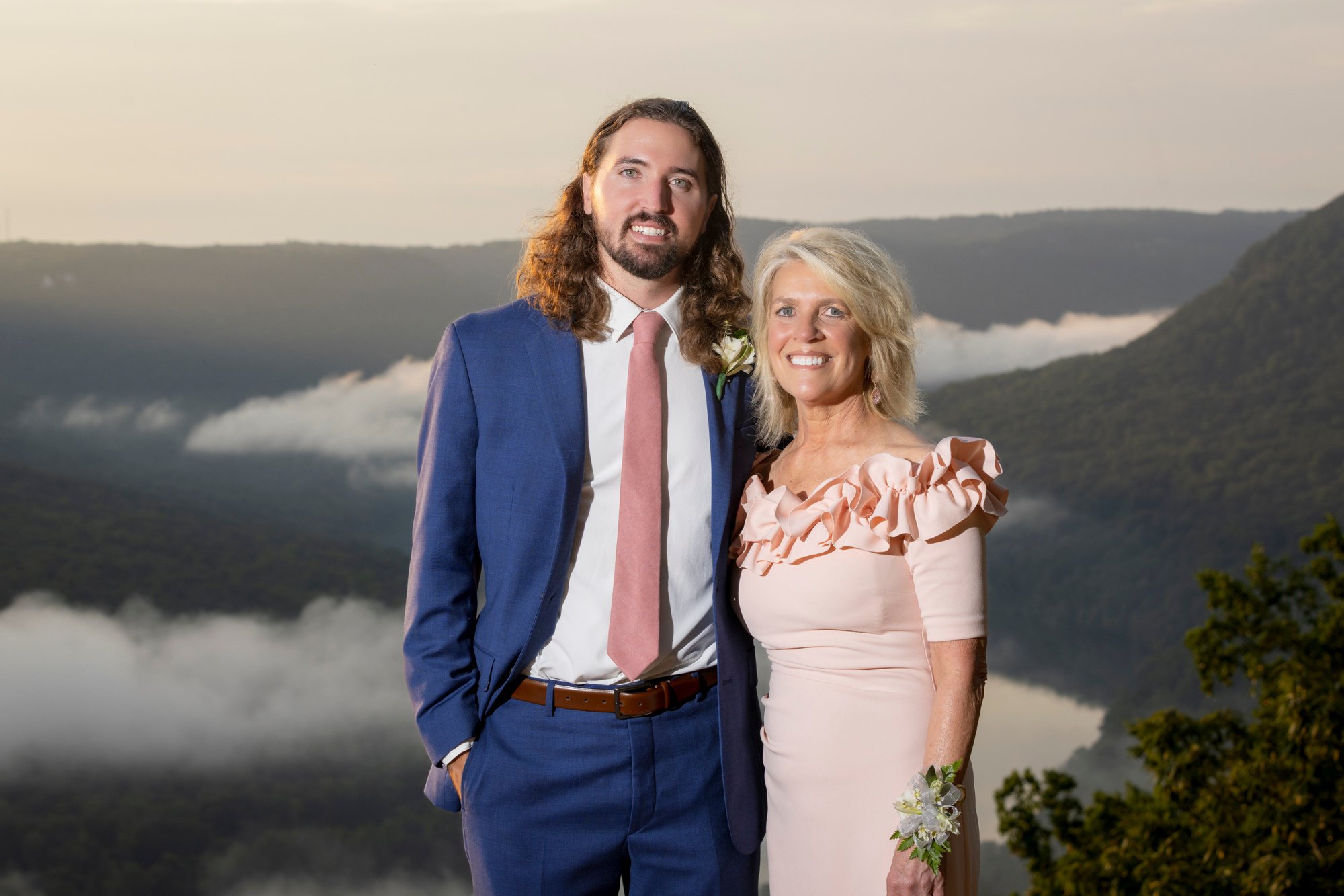 A man in a blue suit and a woman in a light pink dress smiling, standing together outdoors with a misty mountainous landscape in the background.