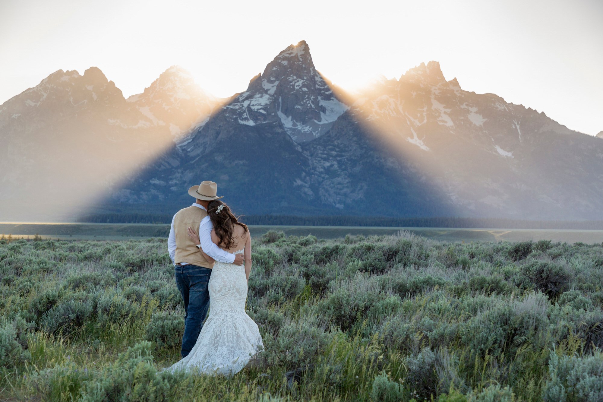 A couple embracing outdoors with mountains and sunlight in the background.