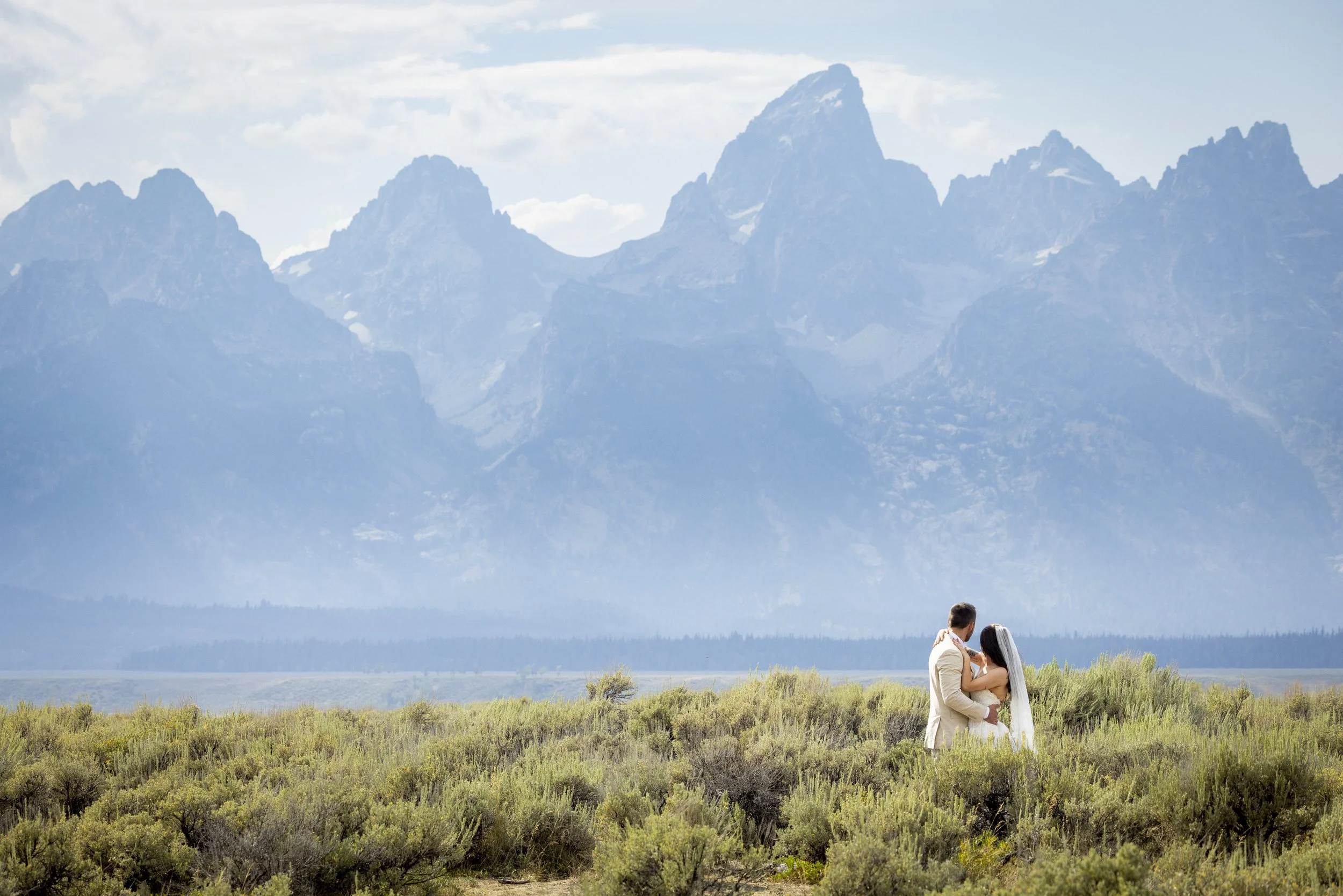 A couple in wedding attire, standing in an open field with mountains in the background, during daytime.