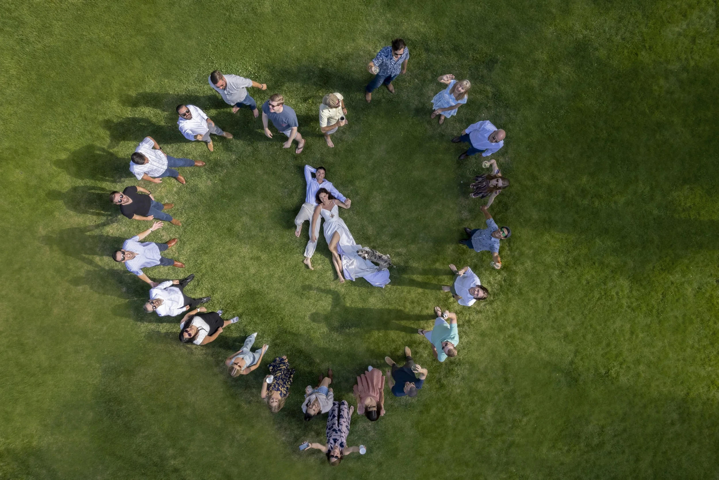 Top-down view of a group of people on a grassy lawn, some lying down and some standing, with a bride and groom lying in the center.