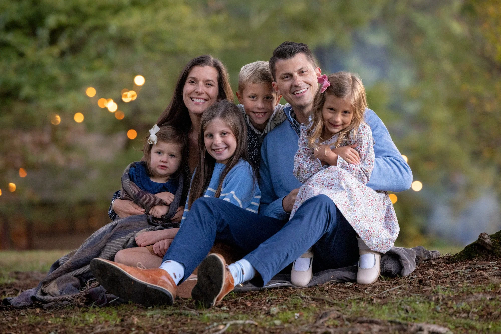 Family of seven sitting and lying on a blanket outdoors in a park with trees and bokeh lights in the background. The family includes two adult men and women, and three young girls and boys. Everyone is smiling.