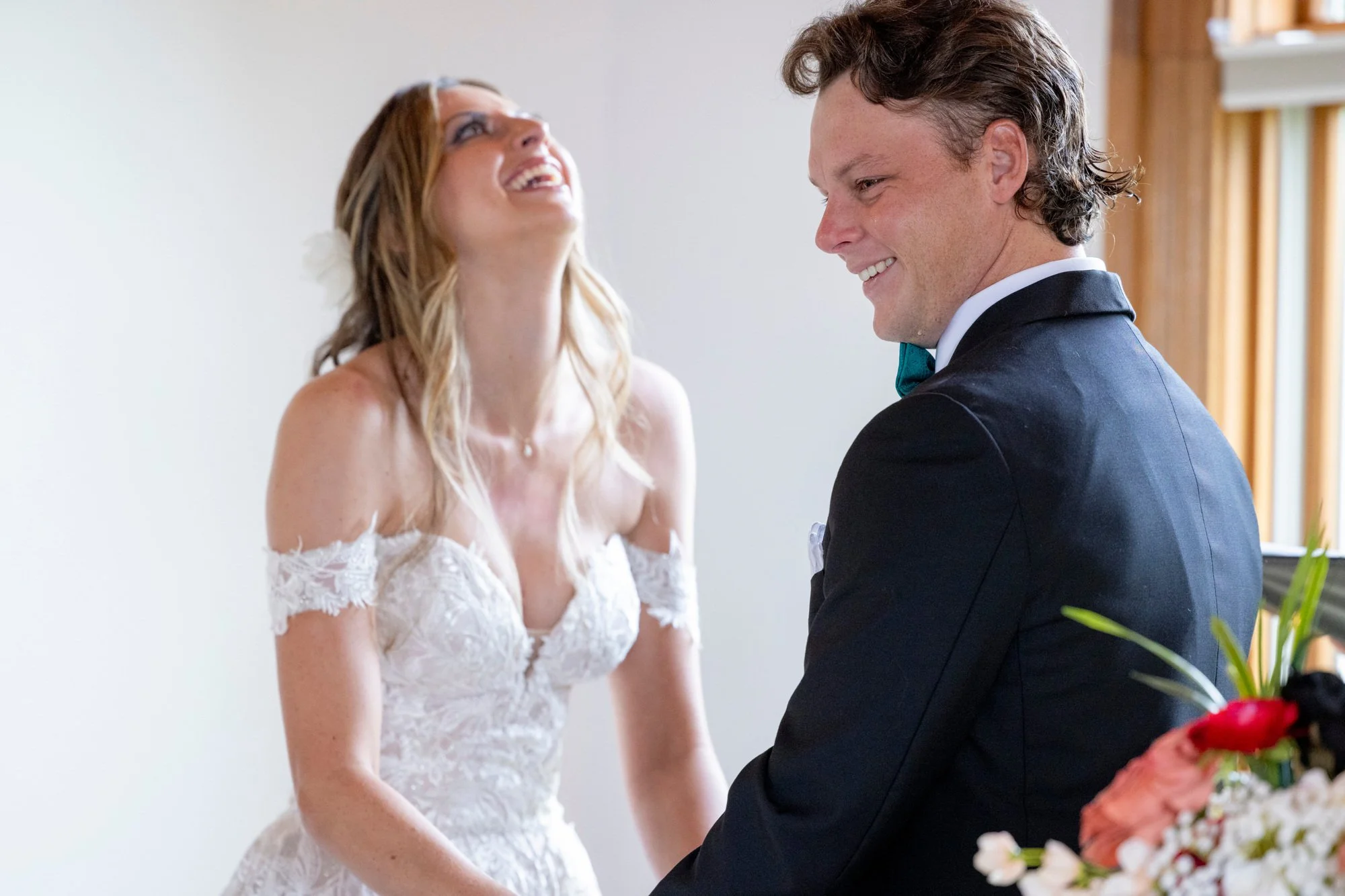 A couple appears to be at a wedding, the woman is wearing a white lace wedding dress and smiling, while the man is wearing a black tuxedo with a bow tie, also smiling. They are facing each other, indoors with a light-colored wall and wooden window bl