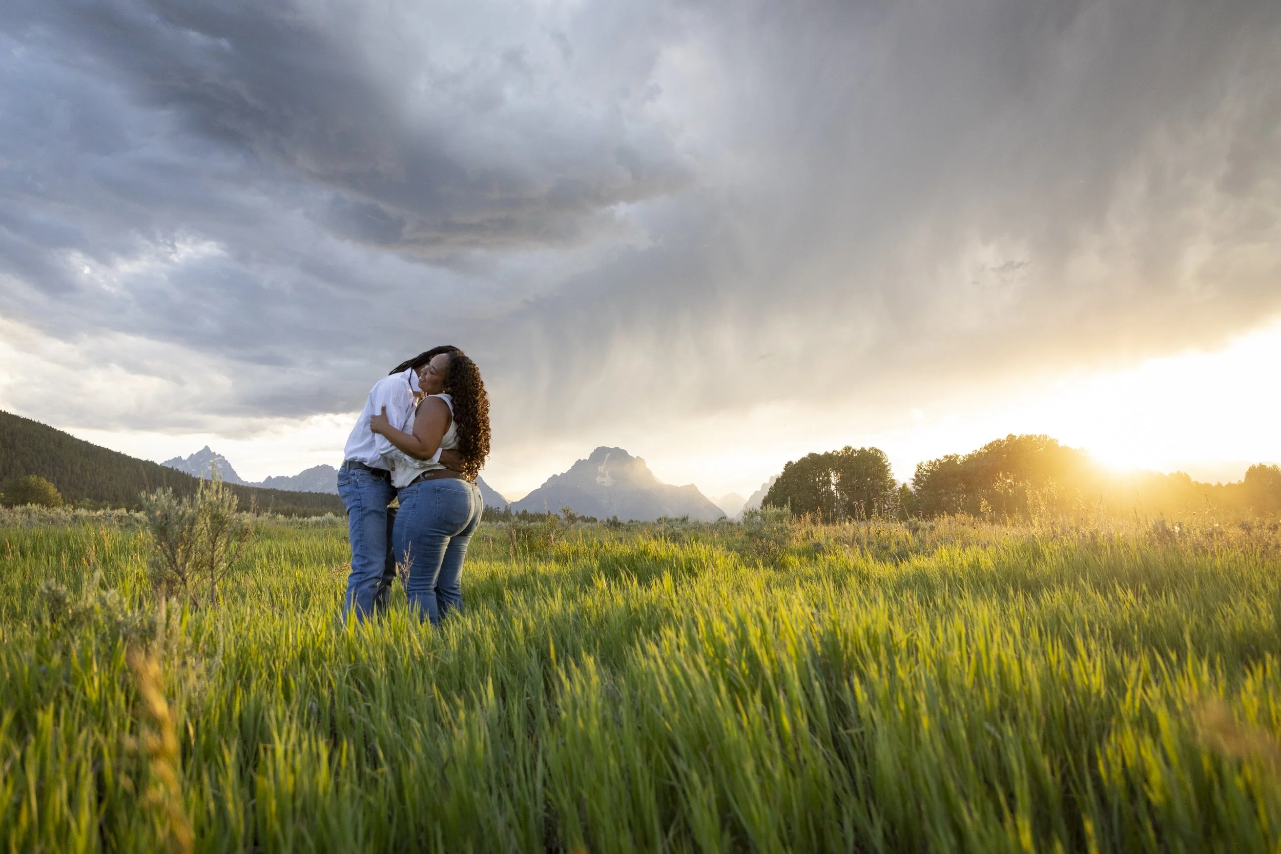 Two women sharing a hug in a grassy field during sunset, with mountains and cloudy sky in the background.