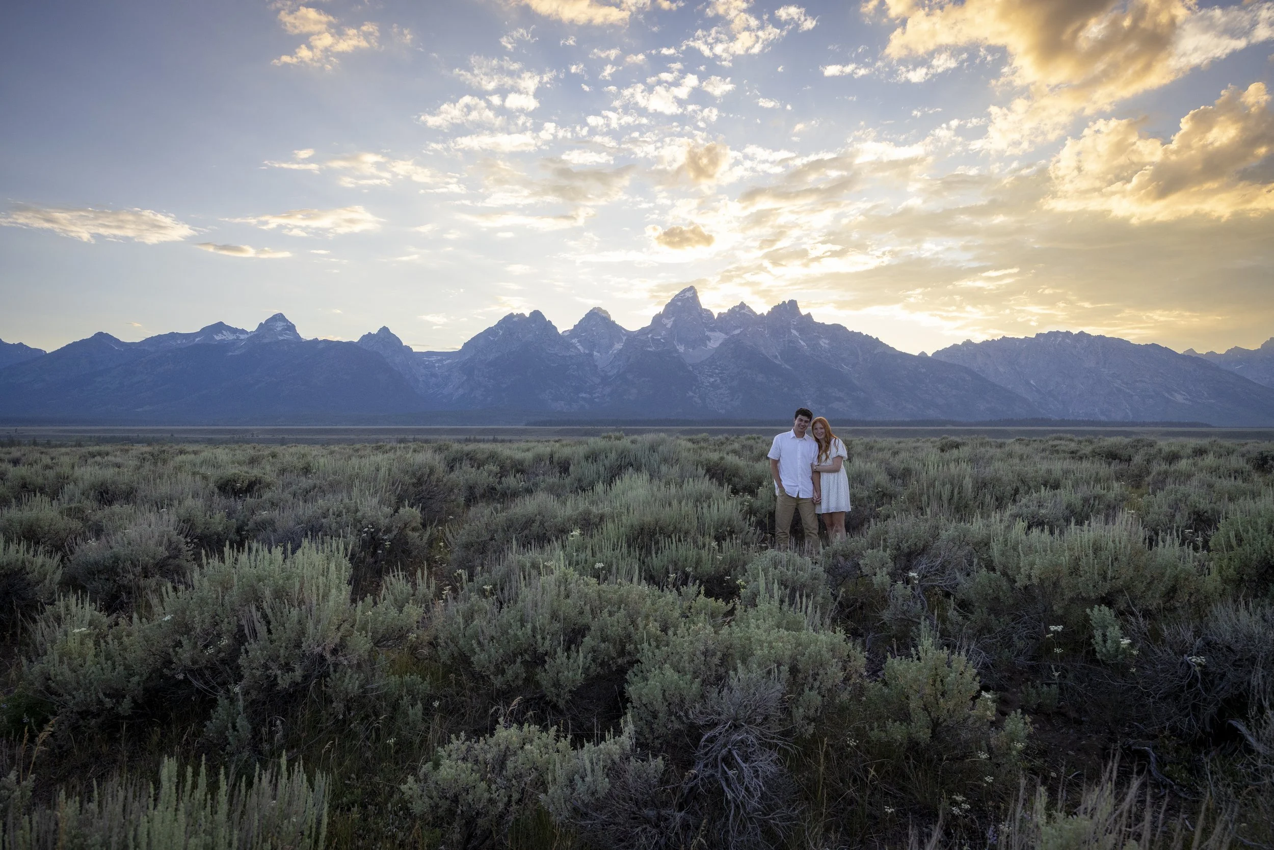 A man and woman standing together in a field of green shrubs with tall mountains and a partly cloudy sky at sunset in the background.